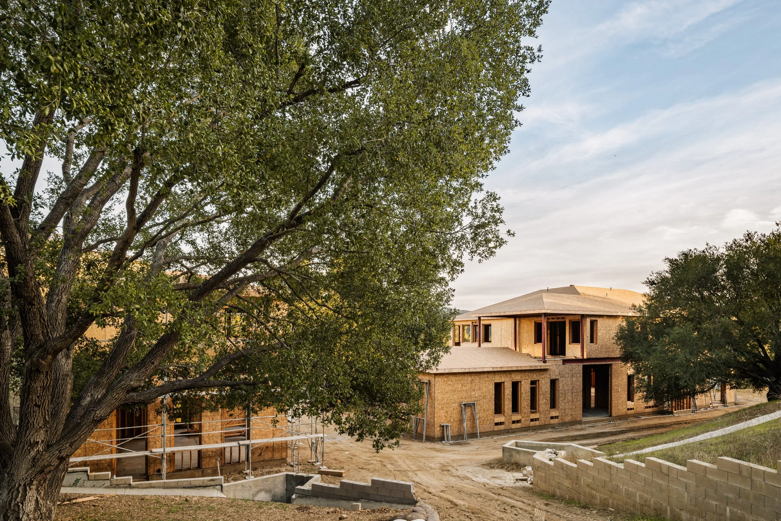 Under construction two-story house with wooden framing, surrounded by trees and a dirt yard.