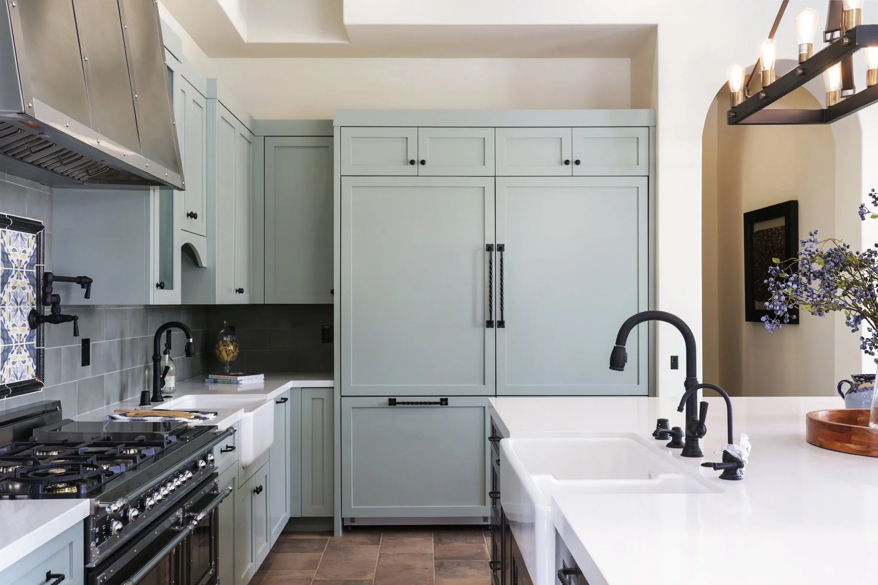 A modern kitchen with light blue cabinets, a white countertop, a black faucet, and a stainless steel stove. There's a large cabinet in the center, a dish soap bottle on the counter, and a decorative jar. The kitchen has wood flooring, black hardware 