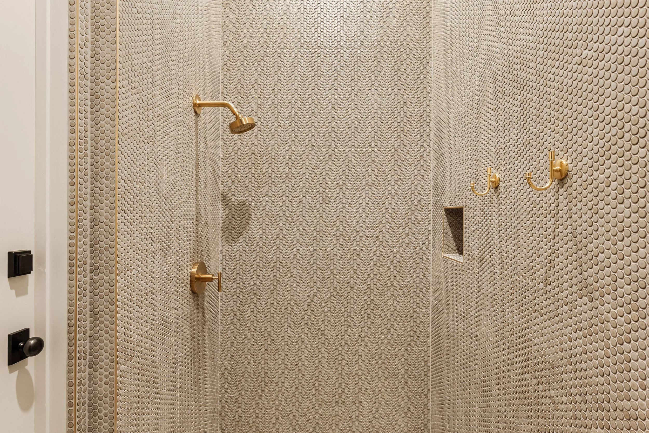 A shower with pebble tile walls, three gold hooks, a gold showerhead, and a small recessed shelf.