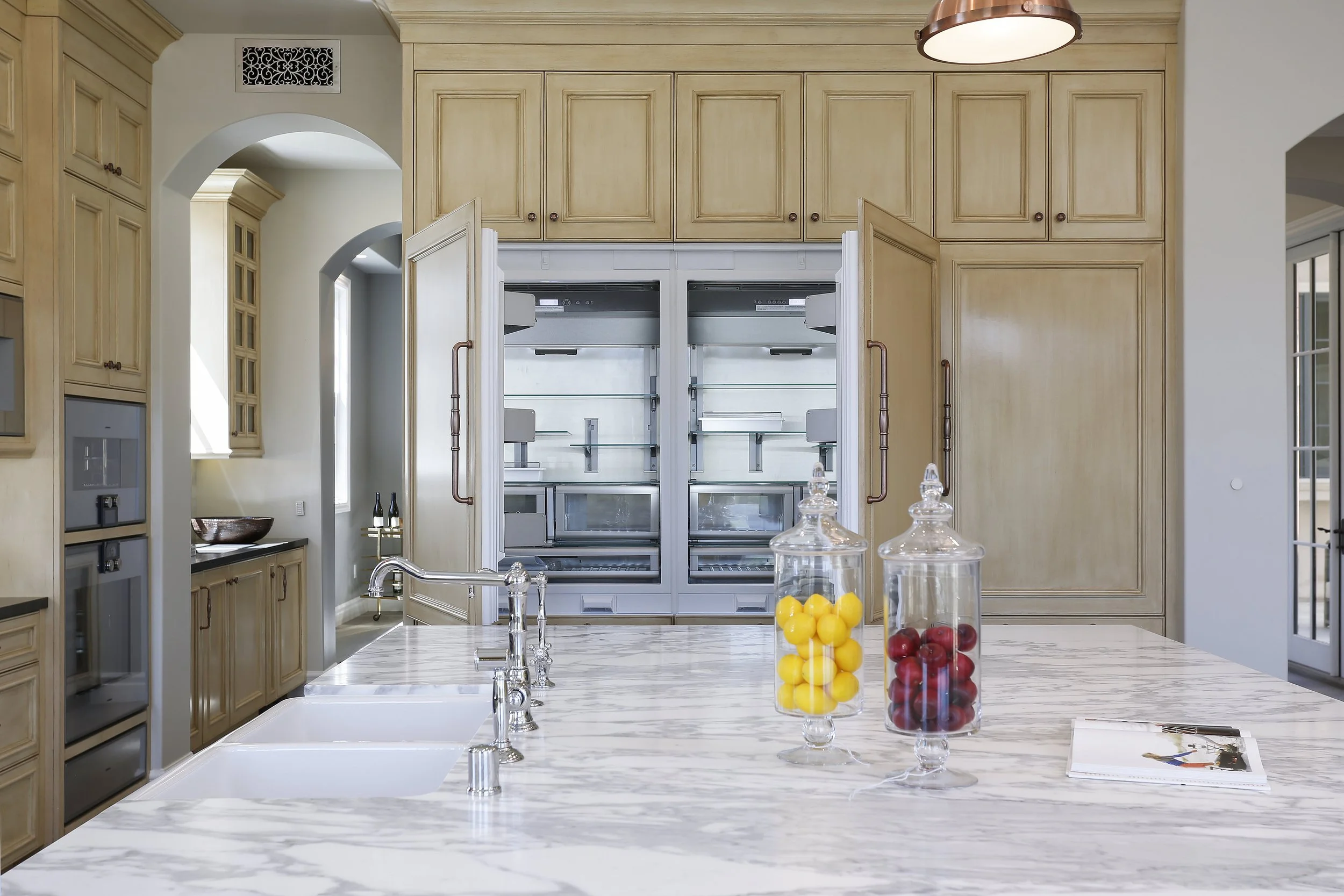 Modern kitchen with beige cabinets and marble countertop island. Two glass jars filled with yellow and red berries sit on the island. Open double oven and a coffee station are visible in the background.