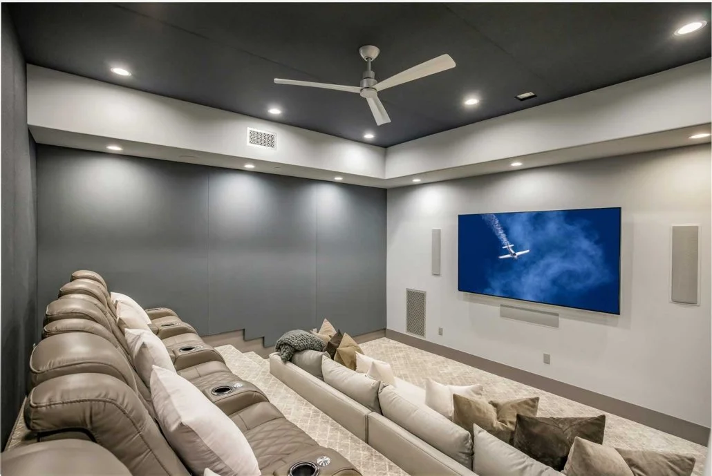A home theater room with beige reclining chairs, a large flat-screen TV on the white wall, beige and brown throw pillows, and a ceiling fan with white blades.
