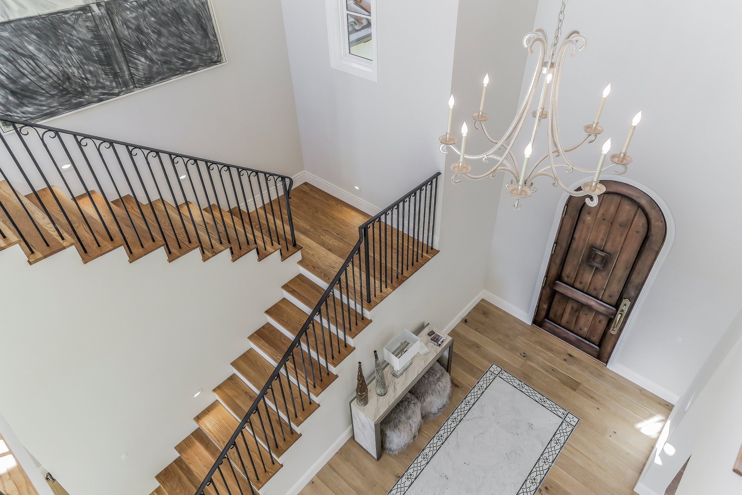 Interior view of a home's entrance foyer with a wooden front door, a chandelier hanging from the ceiling, and a staircase with black railing and wooden steps. There is a console table below the staircase with decorative items, a furry stool, and a pa