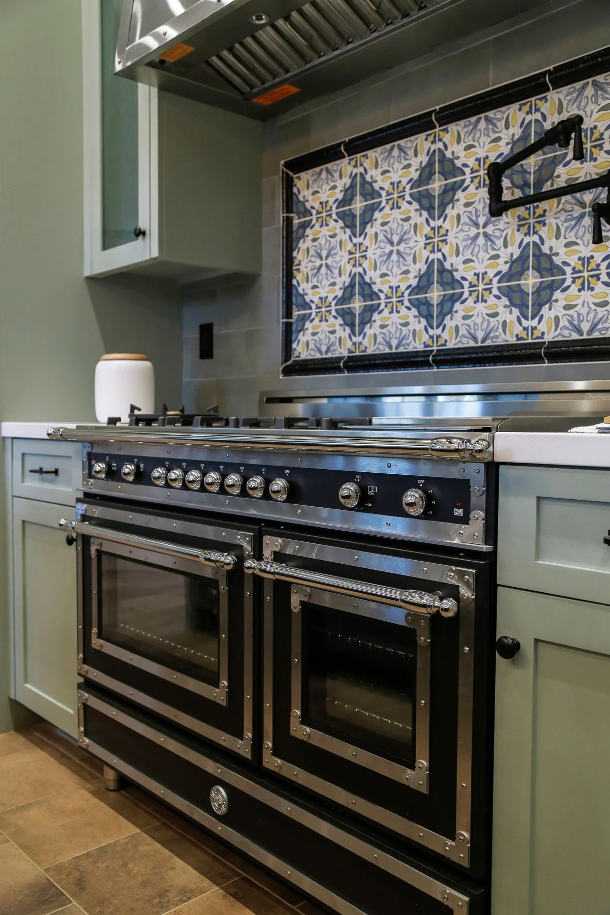 Modern kitchen stove with two ovens, stainless steel finish, against a wall with blue and yellow patterned tile backsplash, green cabinetry, and a white container on the counter.