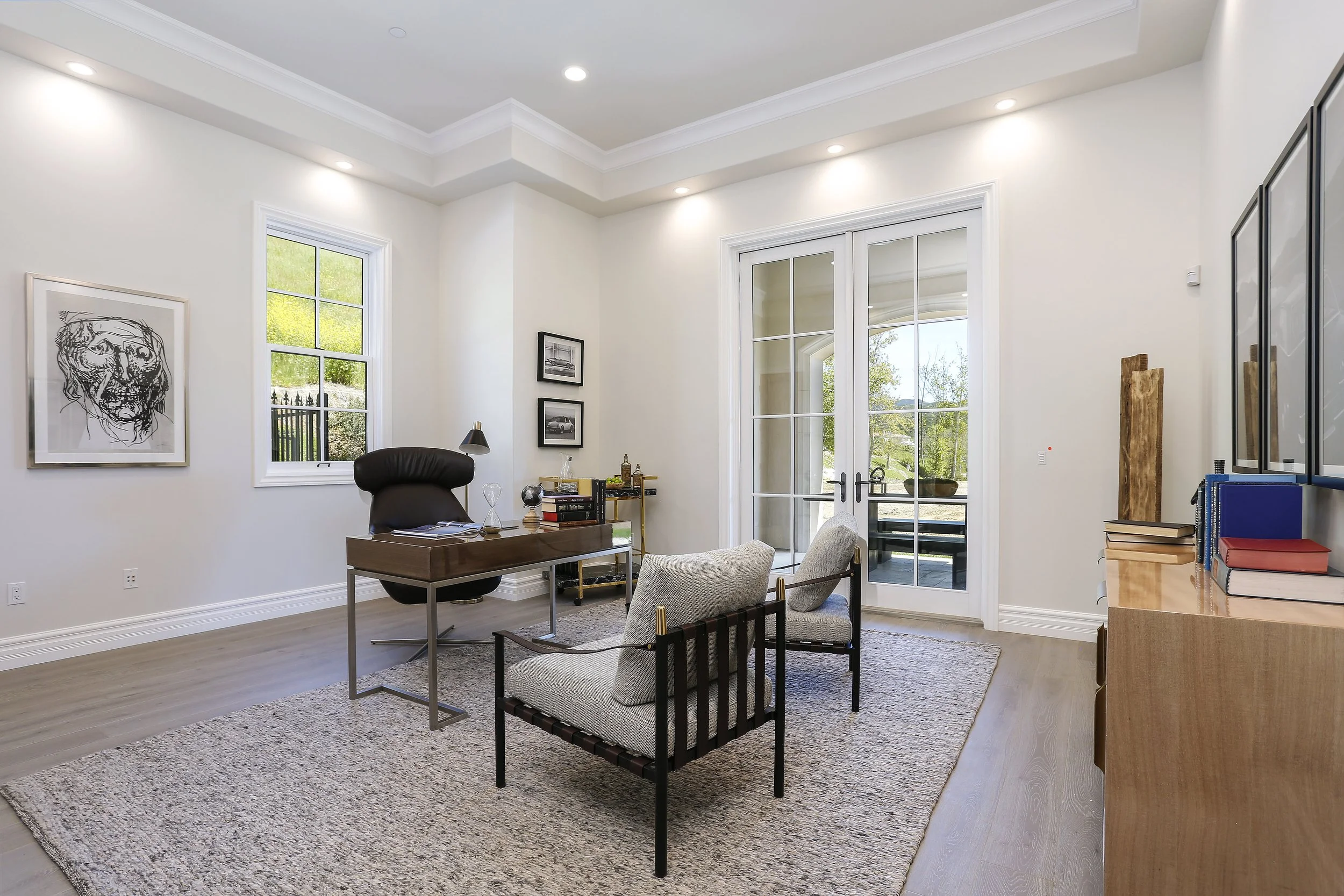 A modern office with white walls, a wooden desk, an office chair, a small sofa, a rug, and a glass door leading to a patio with greenery outside.