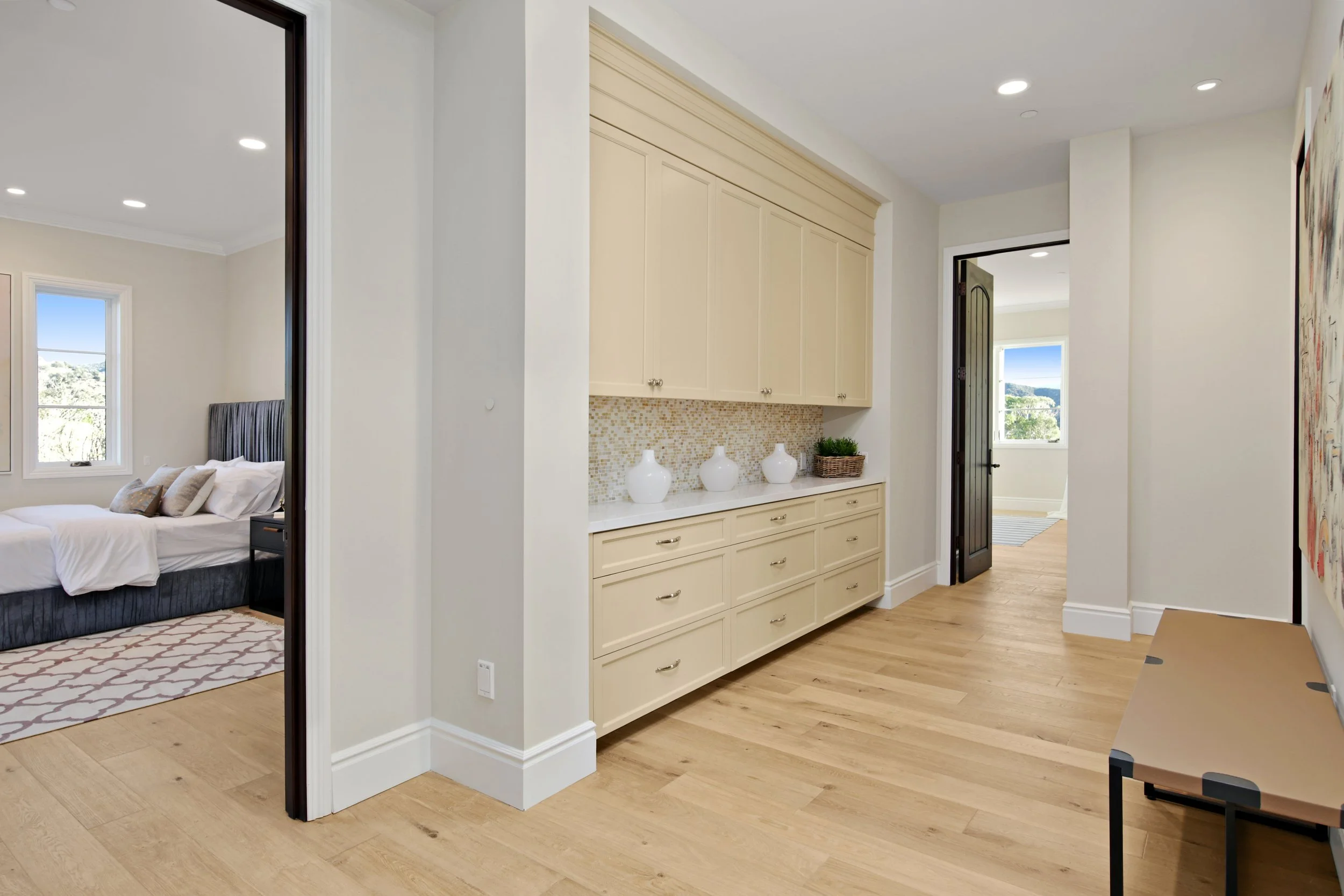 Interior hallway with a cream-colored built-in cabinet, decorative vases, and a basket of plants, with views into bedrooms with large windows showing scenic outdoor views.