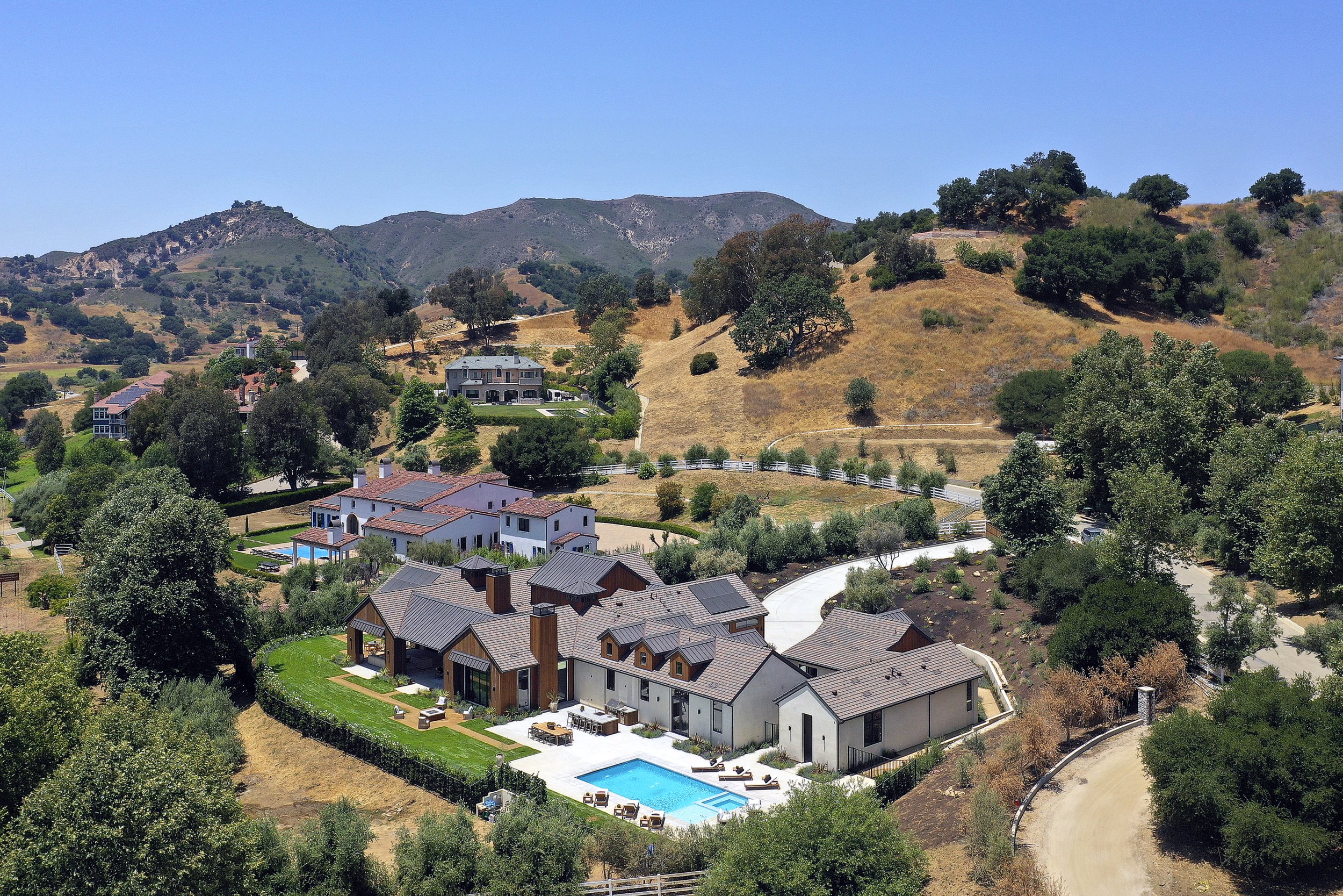 Aerial view of luxurious homes with swimming pools situated on a hillside with trees and dry grass, surrounded by mountains under a clear blue sky.
