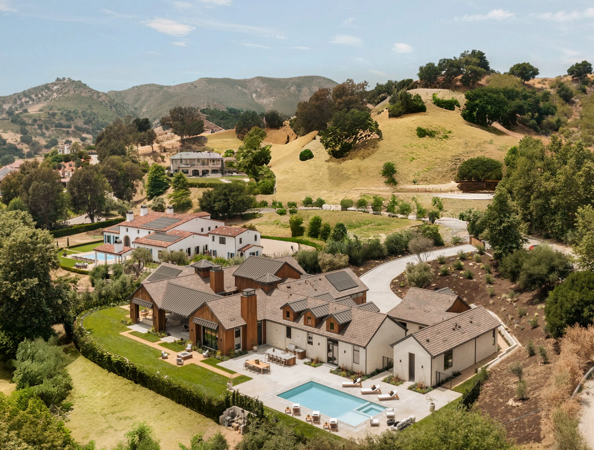 Aerial view of a modern house with a swimming pool, situated in a hilly landscape with other homes, trees, and winding roads.