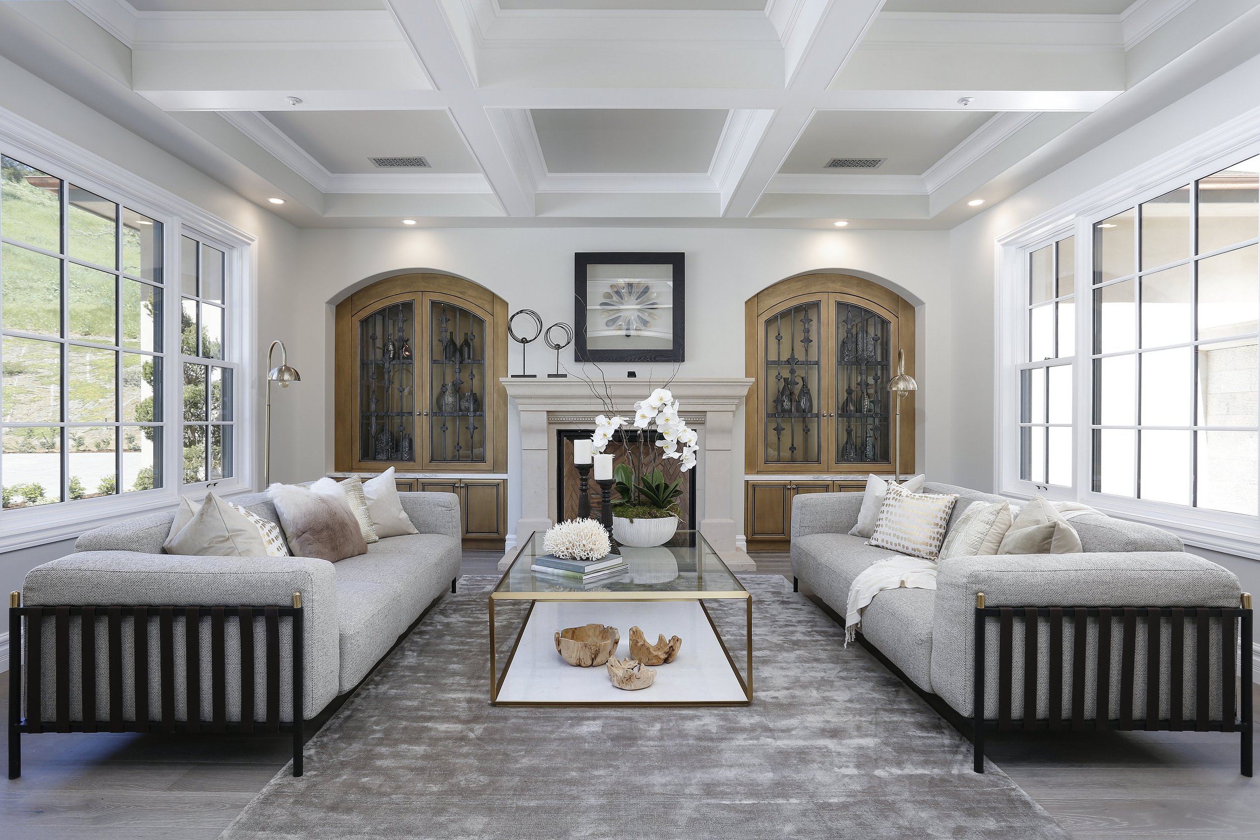 Elegant living room with large windows, white walls, and a coffered ceiling. Two gray sofas face each other with decorative pillows. A glass coffee table with gold frame holds a white orchid plant and decorative items. Built-in wooden cabinets with g