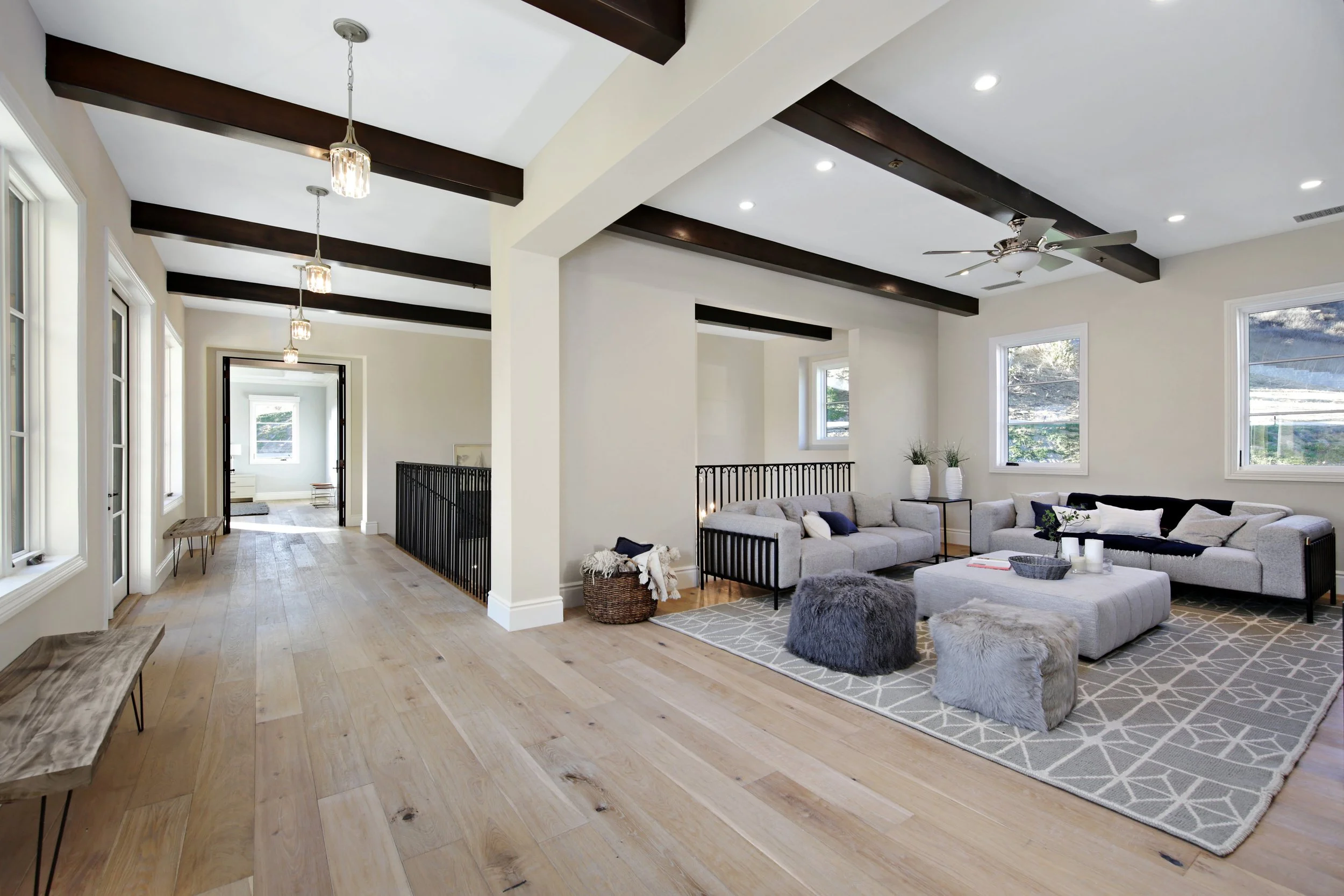 Bright living room with white walls, hardwood floors, black ceiling beams, and large windows. Contains gray sofas, black accents, and textured pillows and ottomans.