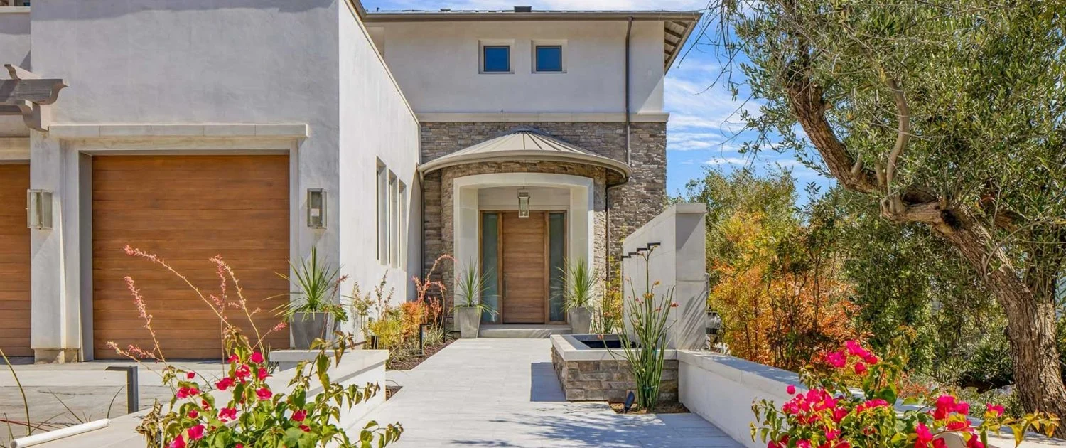 Modern house exterior with wooden garage doors, a stone and stucco entrance, potted plants, colorful flowers, and trees on a sunny day.