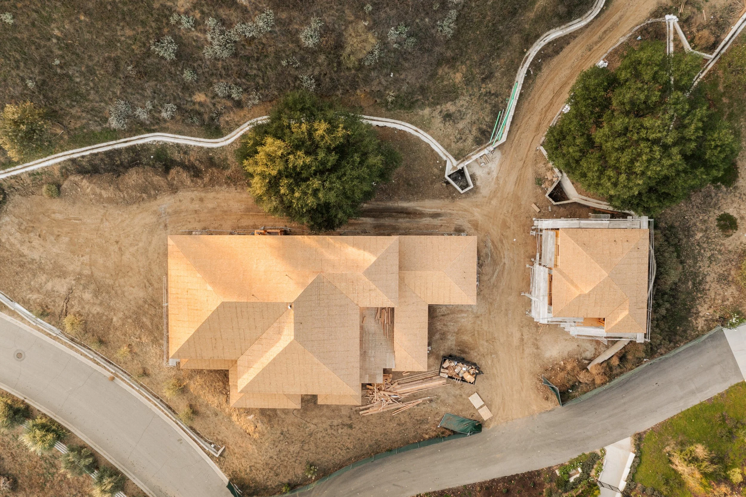 An aerial view of a house under construction with a roof, a dirt driveway, and surrounding trees.