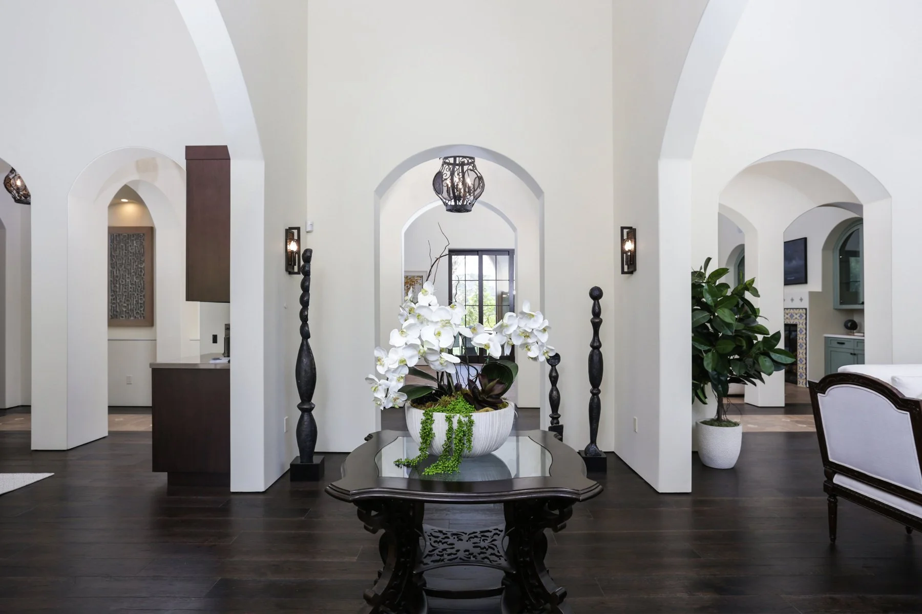 Elegant interior with archways, a dark wooden table with a white orchid plant, and a large potted plant, featuring dark hardwood floors and modern lighting.