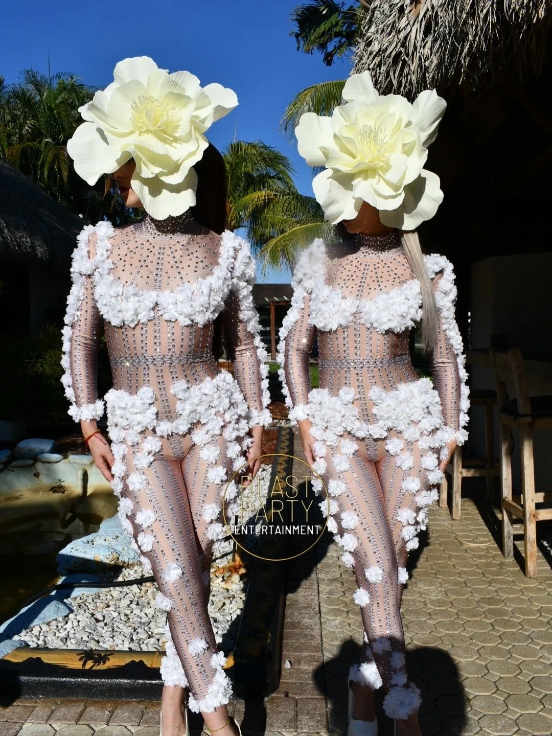 Two women in matching embellished sheer jumpsuits with white floral appliqués, wearing large white flower headpieces, standing outdoors with palm trees and a thatched roof in the background.