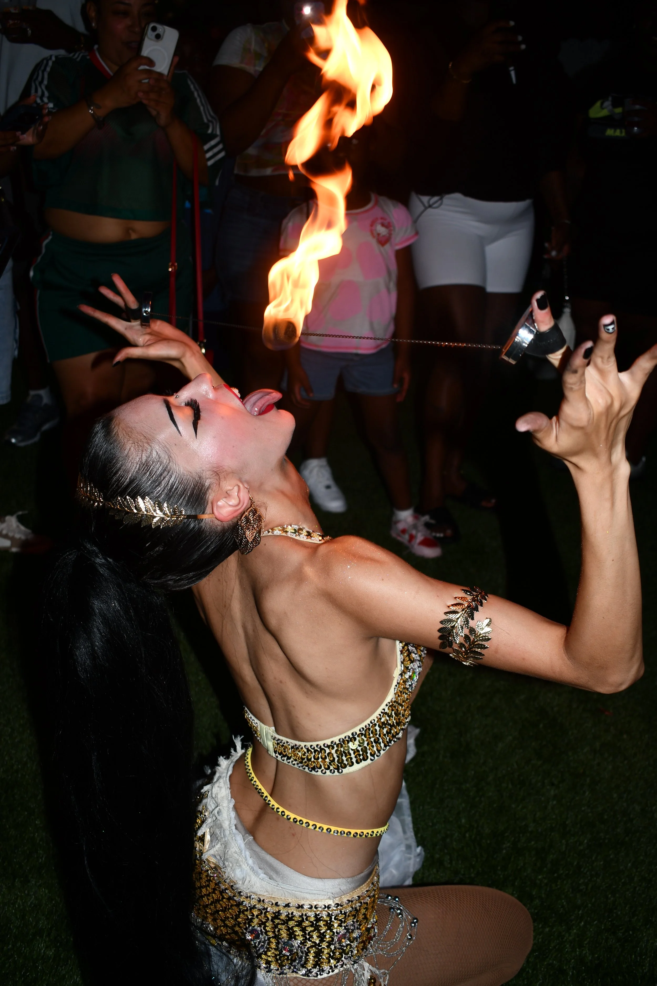 A woman, dressed in an ornate costume with gold and black embellishments and jewelry, is performing a fire-eating act at an event, with her mouth open and a flame close to her face. Spectators, including a child in a pink and white shirt and others taking photos, are watching the performance in the background.