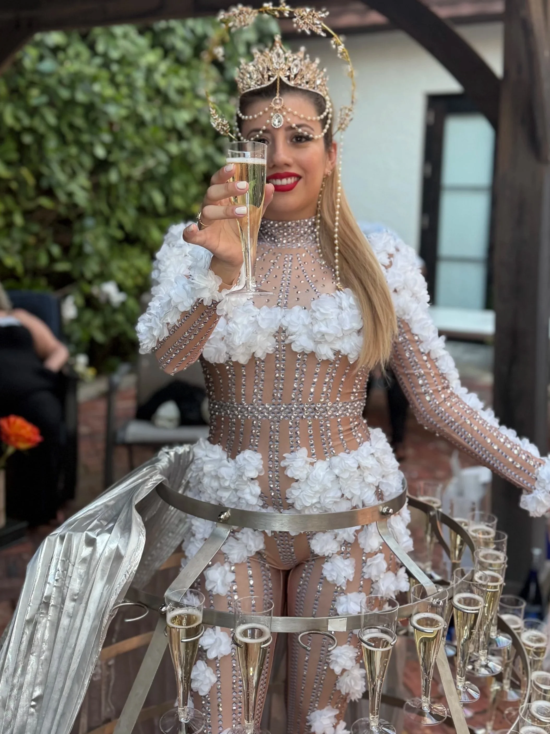 A woman dressed in an ornate, jeweled costume with a crown and pearl accessories, holding a champagne flute, standing behind a table with multiple glasses of champagne in a garden or outdoor setting.