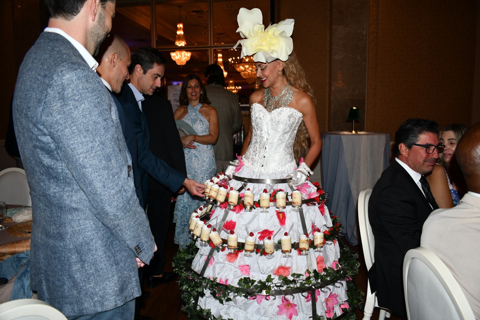 A woman dressed in a white lace corset and skirt with a large floral headpiece stands behind an elaborate tiered display of small dessert glasses at a formal event. Several guests observe and smile as they participate.