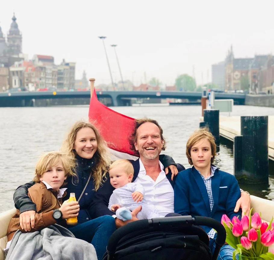 Family sitting in a boat on a canal in an urban area, with historic buildings and a bridge in the background, and pink tulips in the foreground.