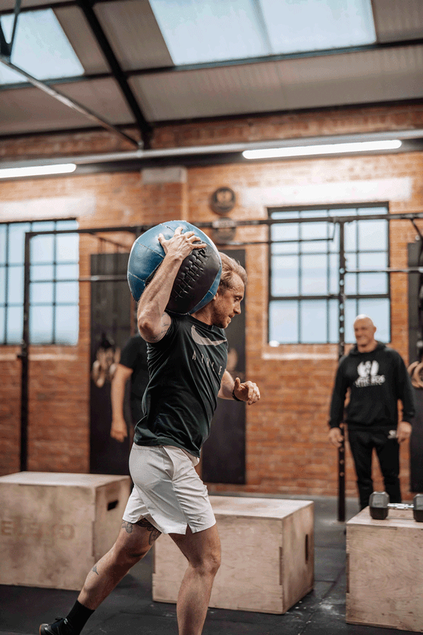 Man lifting a medicine ball in a gym with brick walls and large windows, with two other men watching.