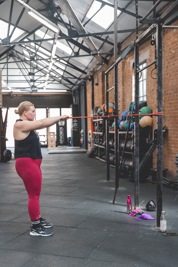 A woman performs a resistance band exercise in a gym with brick walls and large windows, with workout equipment and water bottles nearby.