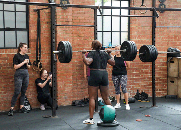 A woman with short hair lifts a barbell with weights in a gym. Several women watch and take photos against brick walls, gym equipment and windows are visible.