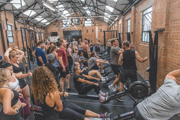 Indoor fitness class with people using rowing machines, some waiting, in a brick-walled gym with large windows and workout equipment.