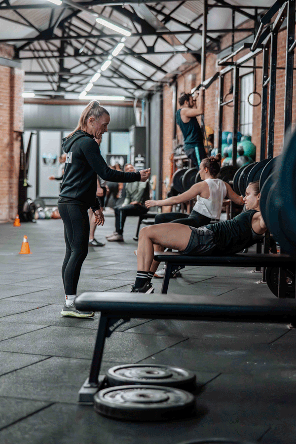 People working out at a gym with exercise equipment and weights, with trainer guiding one person.