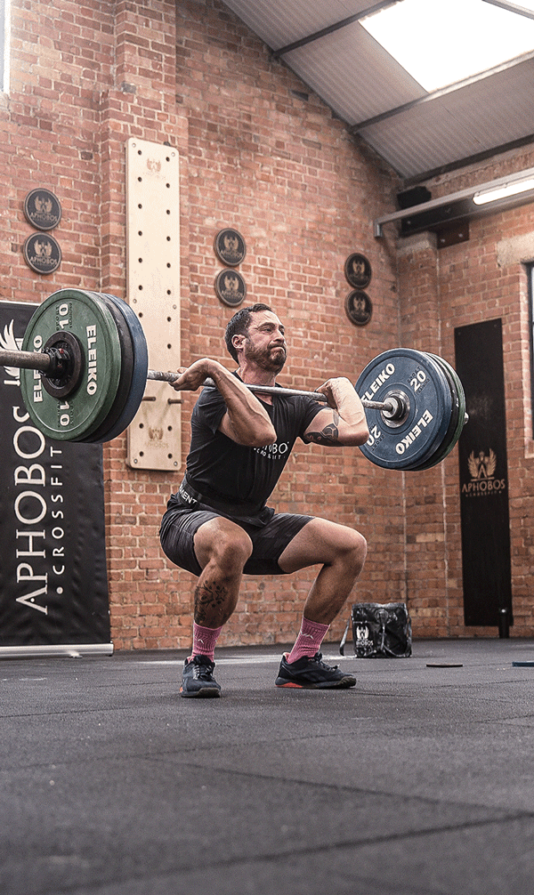 A man wearing a black t-shirt and shorts performing a squat with a loaded barbell at a gym with brick walls and a black banner.