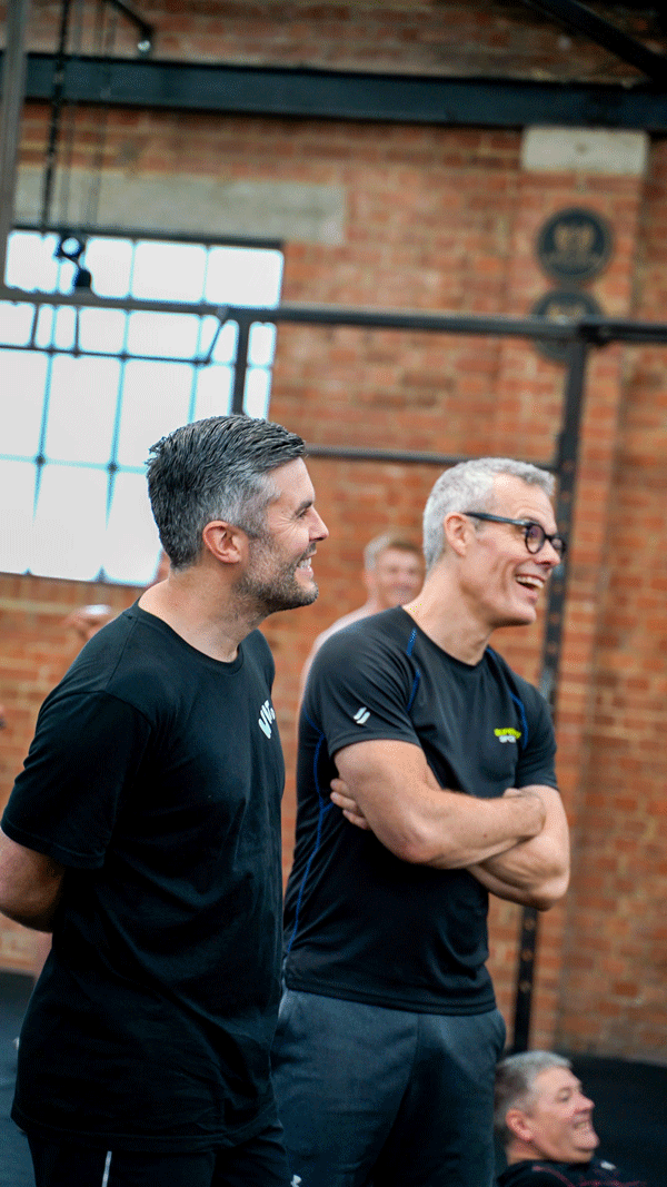 Two men are standing inside a gym, smiling and engaging with each other. One has dark hair and a beard, the other has gray hair and glasses. The gym has exposed brick walls and large windows.