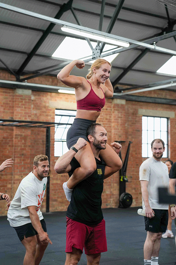 A group of people in a gym celebrating; a woman in a red sports bra and gray shorts is sitting on a man's shoulders, flexing her arms and smiling, while the man supports her with his hands; three other men are nearby, one smiling and looking at the c