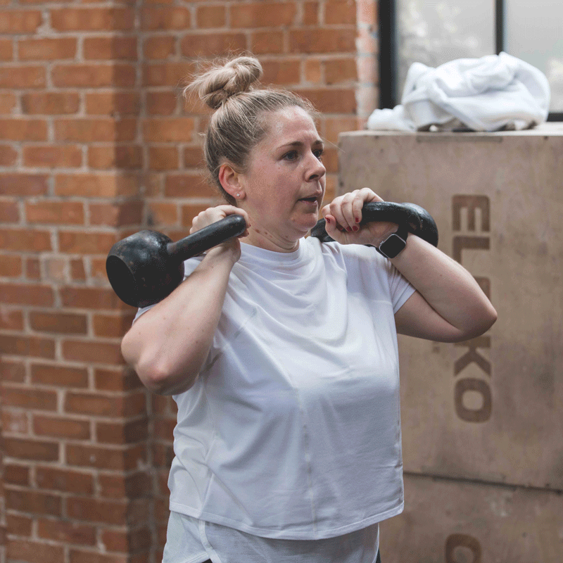 A woman with blonde hair in a bun lifting a kettlebell in a gym with brick walls.