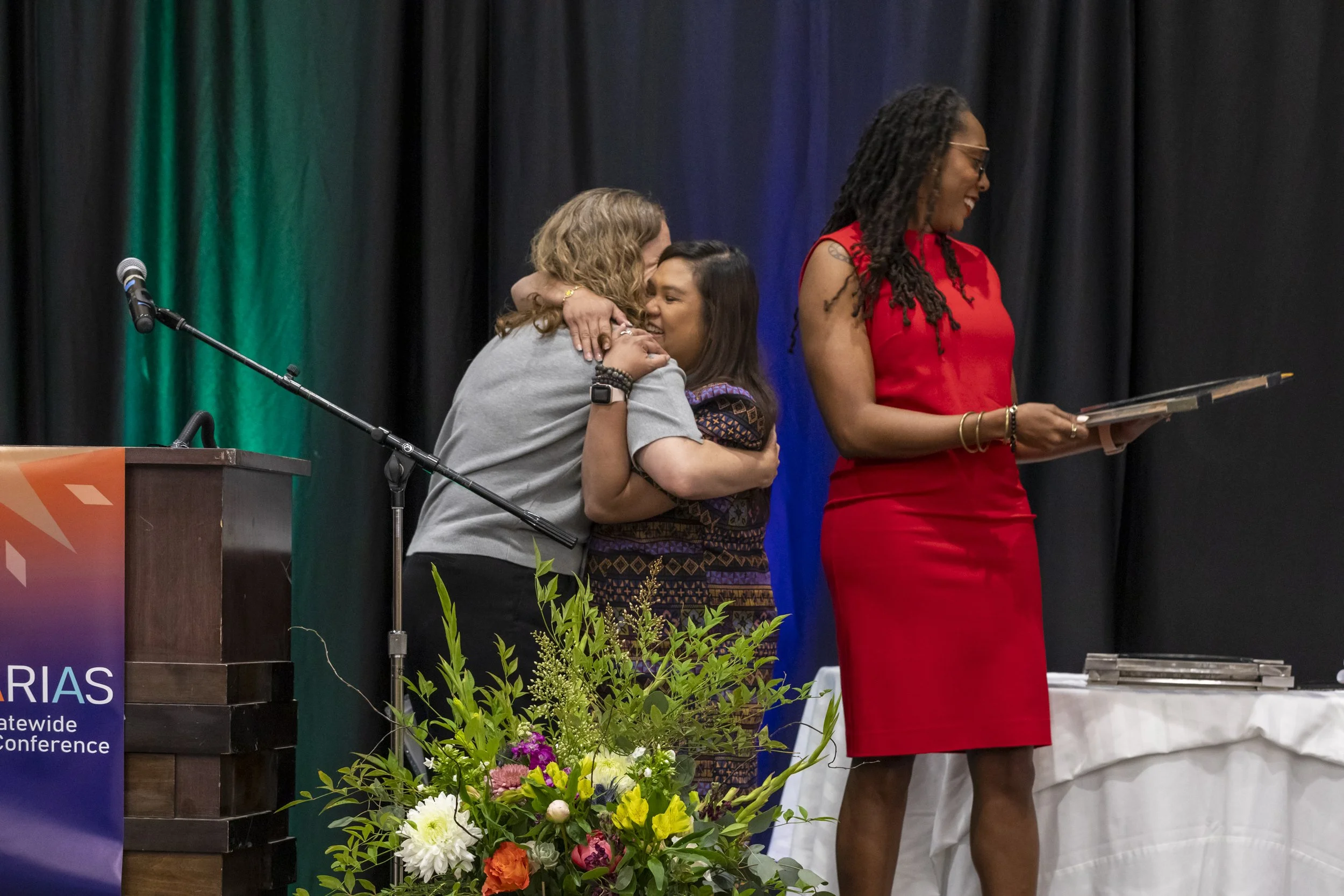 Two women hugging on stage while another holds an award
