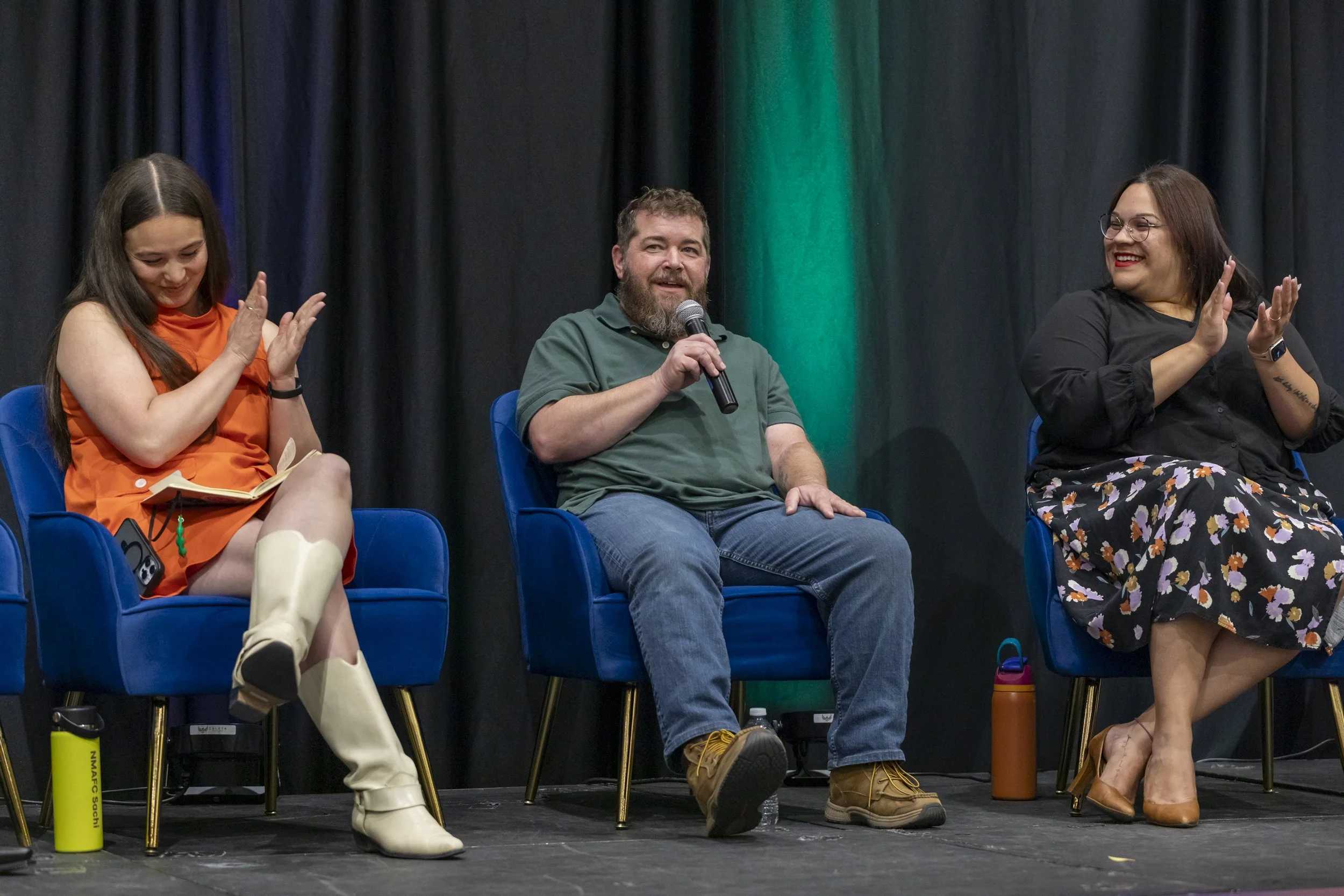 A man speaking into a mic on stage with two other women clapping