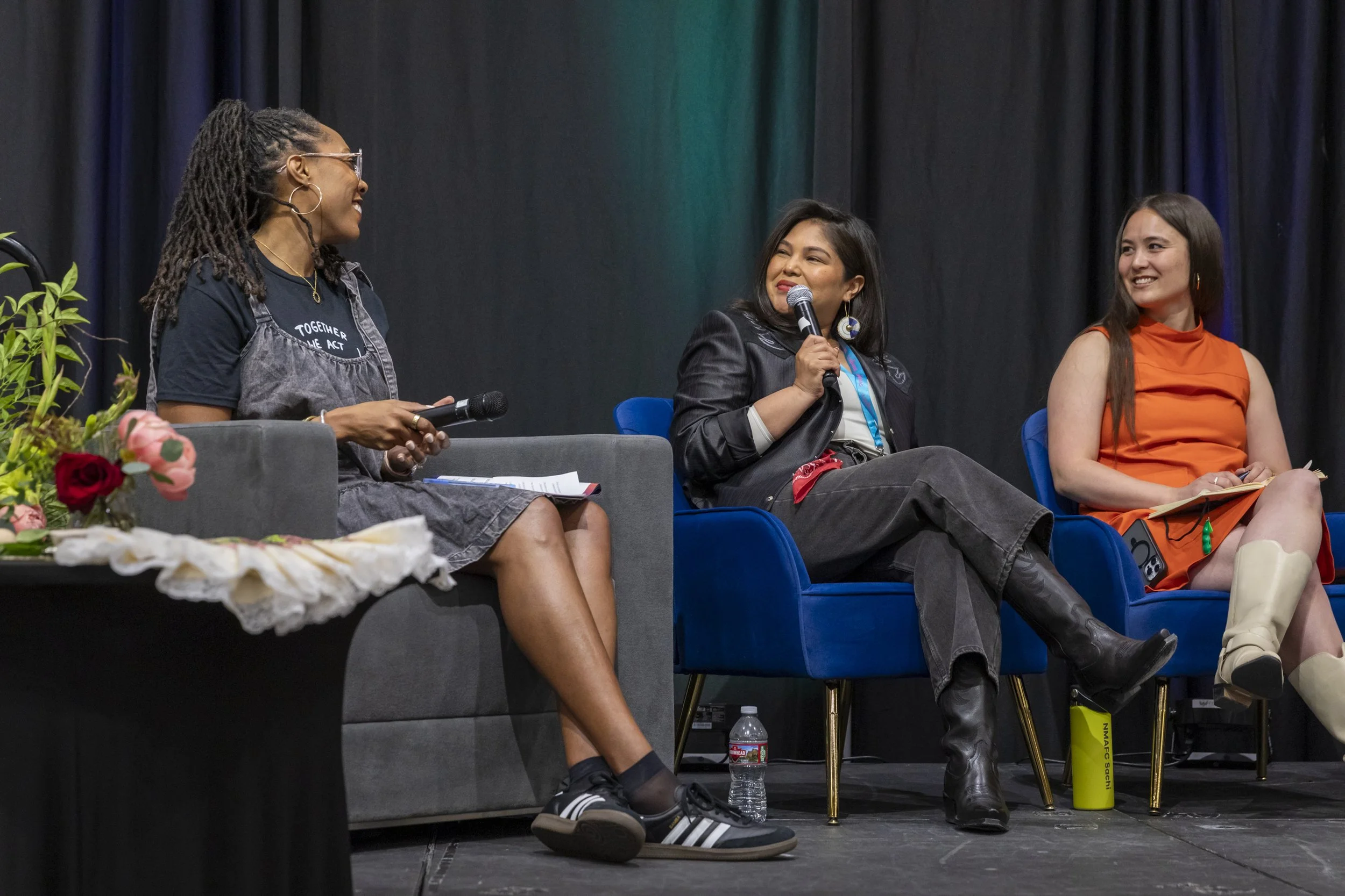 A woman speaking into a mic on stage with two other women