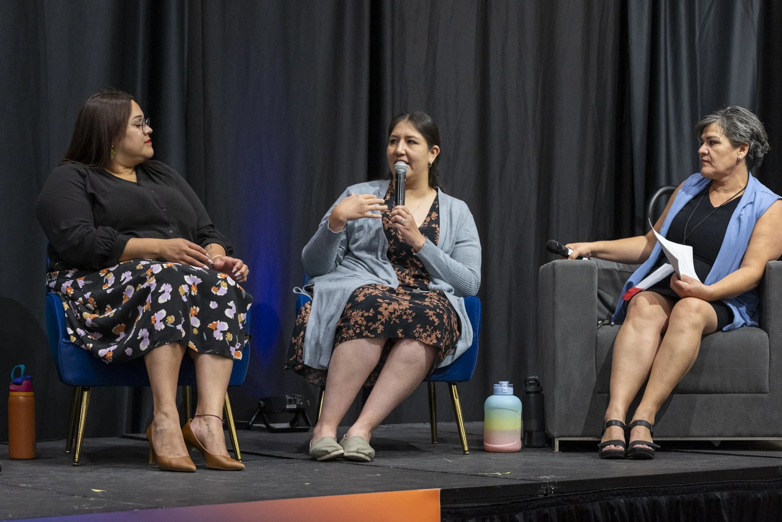 A woman speaking into a mic on stage with two other women