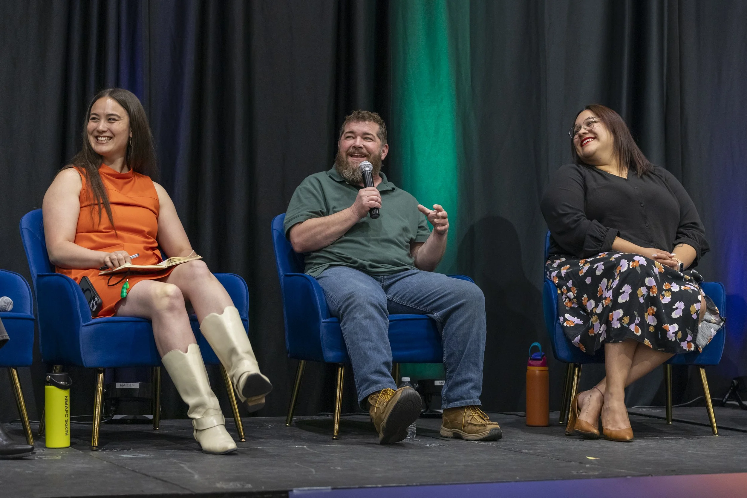 A man speaking into a mic on stage with two other women