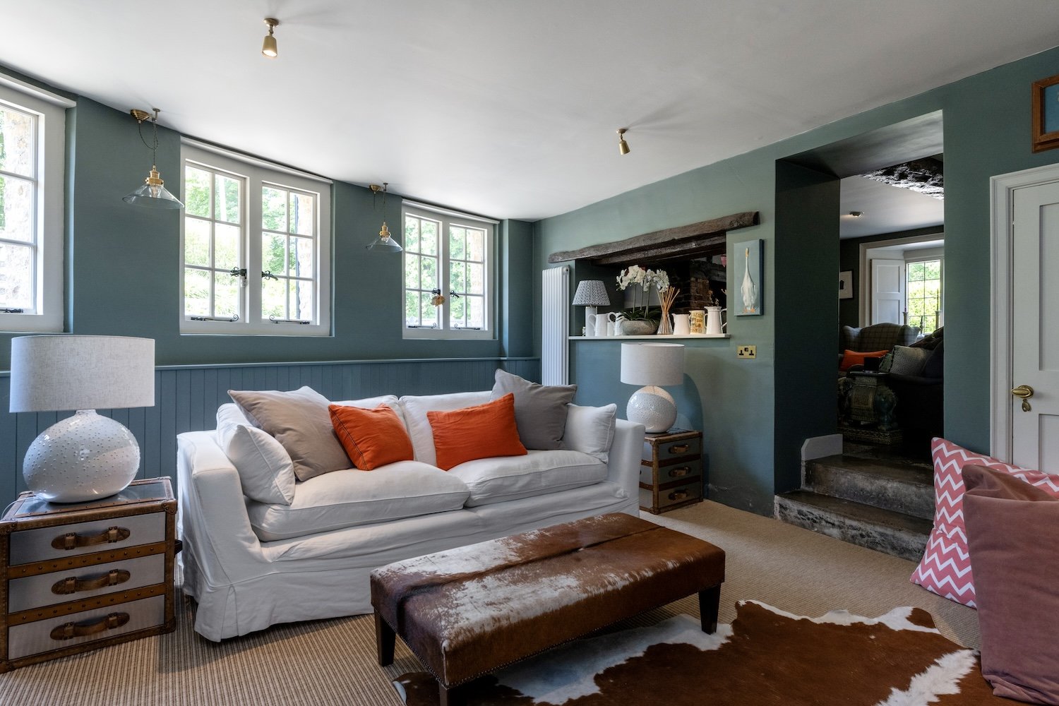 Living room with a white sofa, orange and beige cushions, wooden side tables, lamps, and large windows with greenery outside.