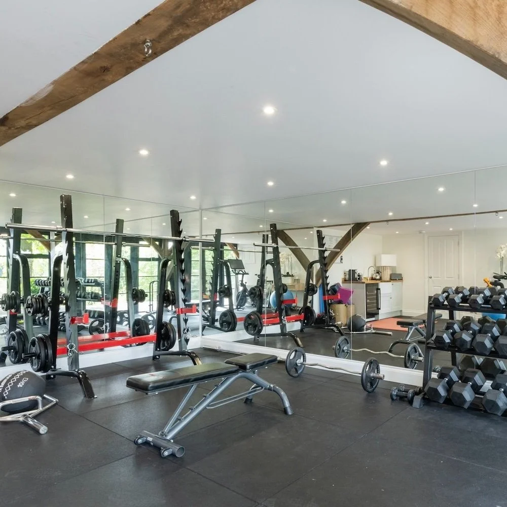 Interior of a gym with a mirror wall, weights and benches and glass doors open on to the garden