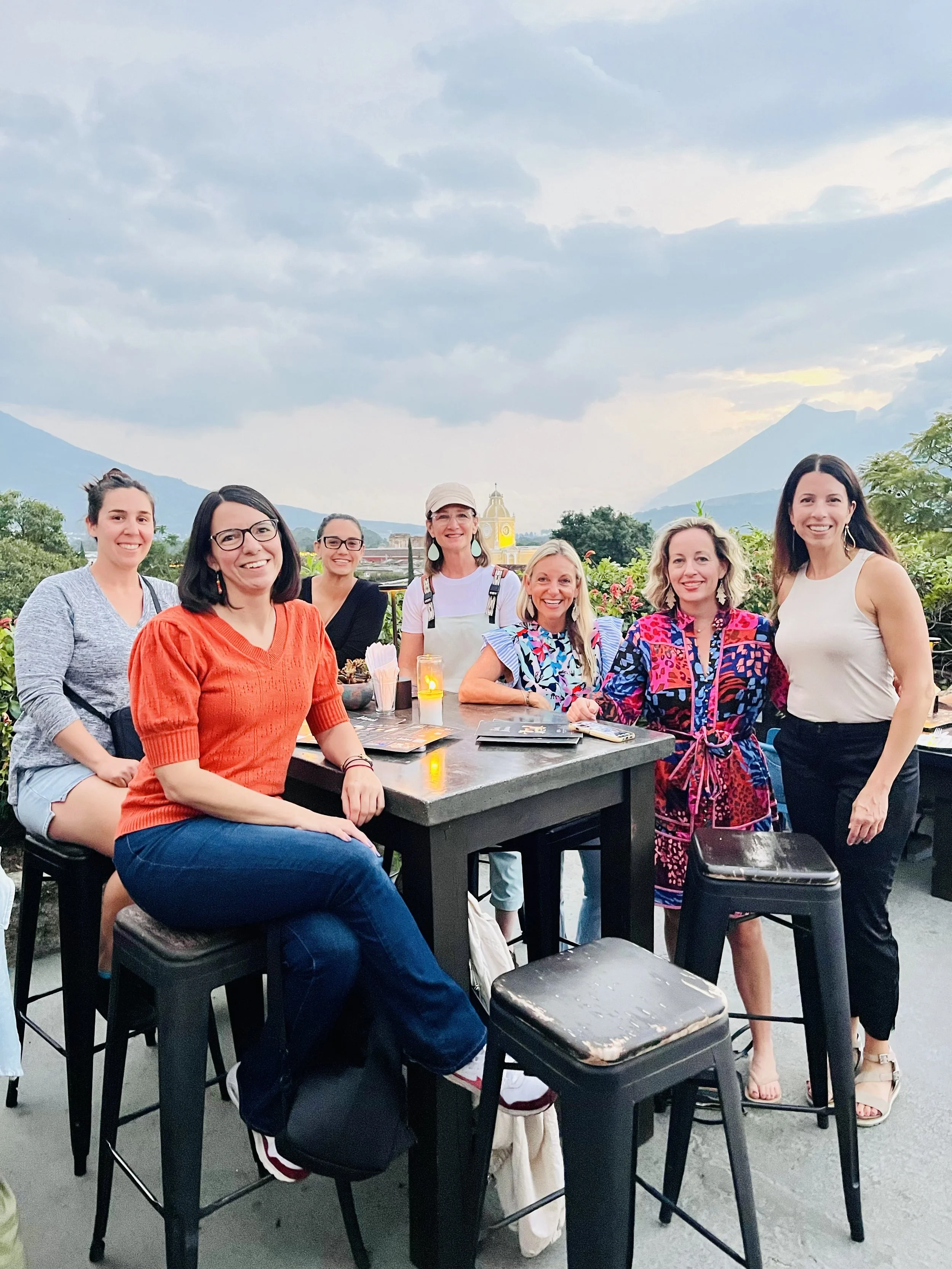 A group of seven women sitting and standing around a dark-colored outdoor table on a rooftop, with a scenic mountain and cloudy sky background, during daytime.