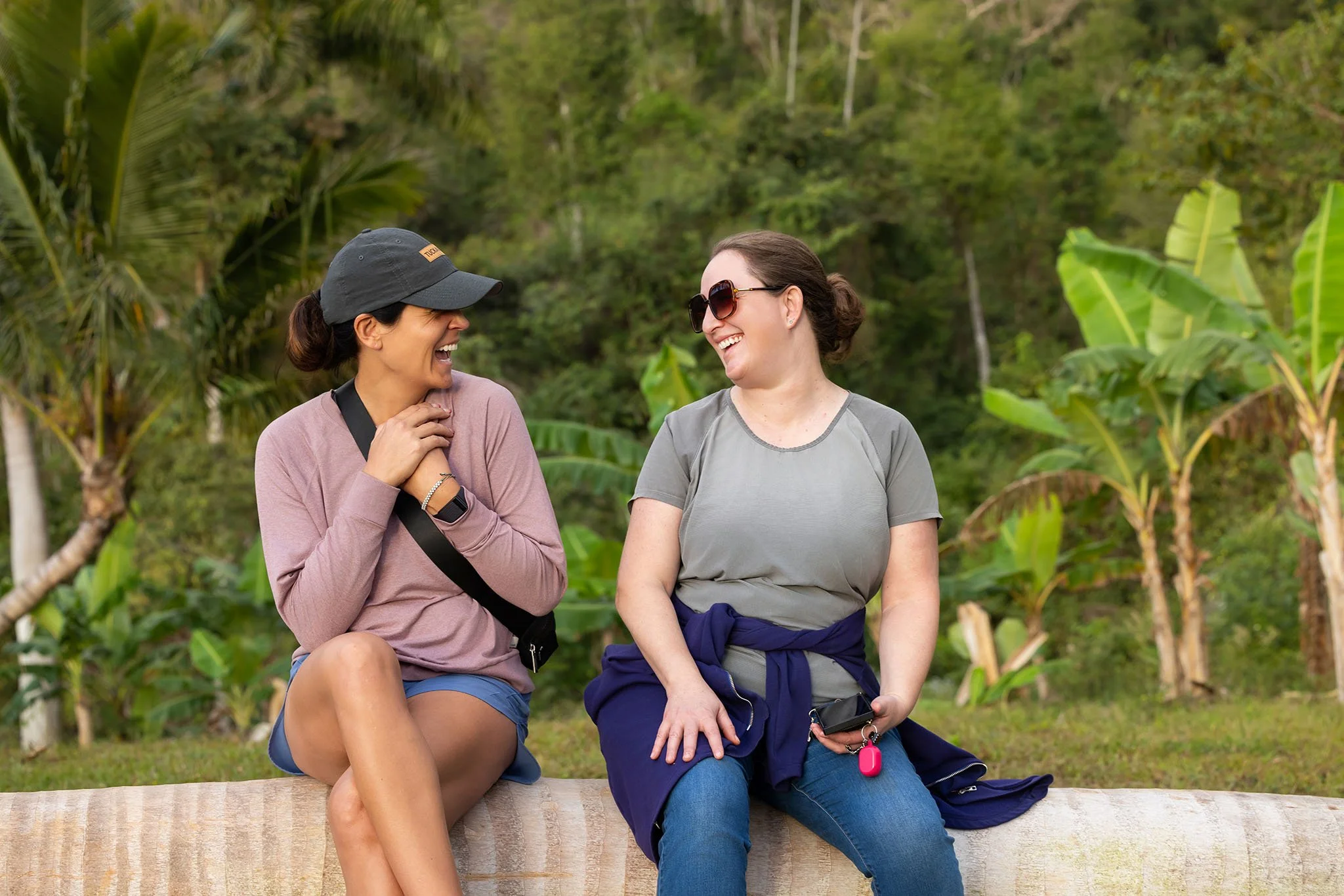 Two women sitting on a log outdoors, laughing and enjoying each other's company, with lush green trees and banana plants in the background.