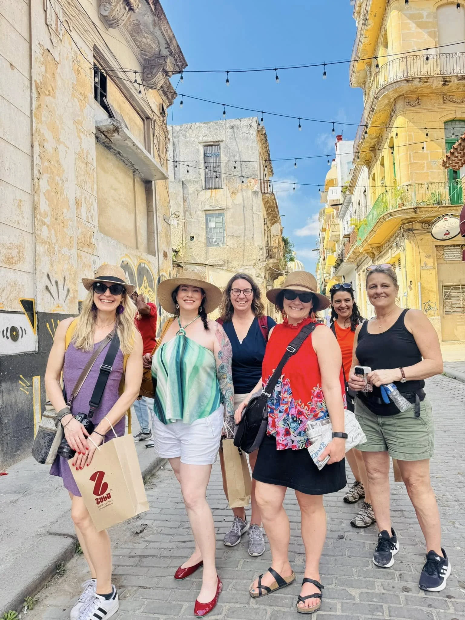 A group of seven women tourists standing on a cobblestone street in a colorful, historic neighborhood with old buildings and string lights, smiling for a photo.