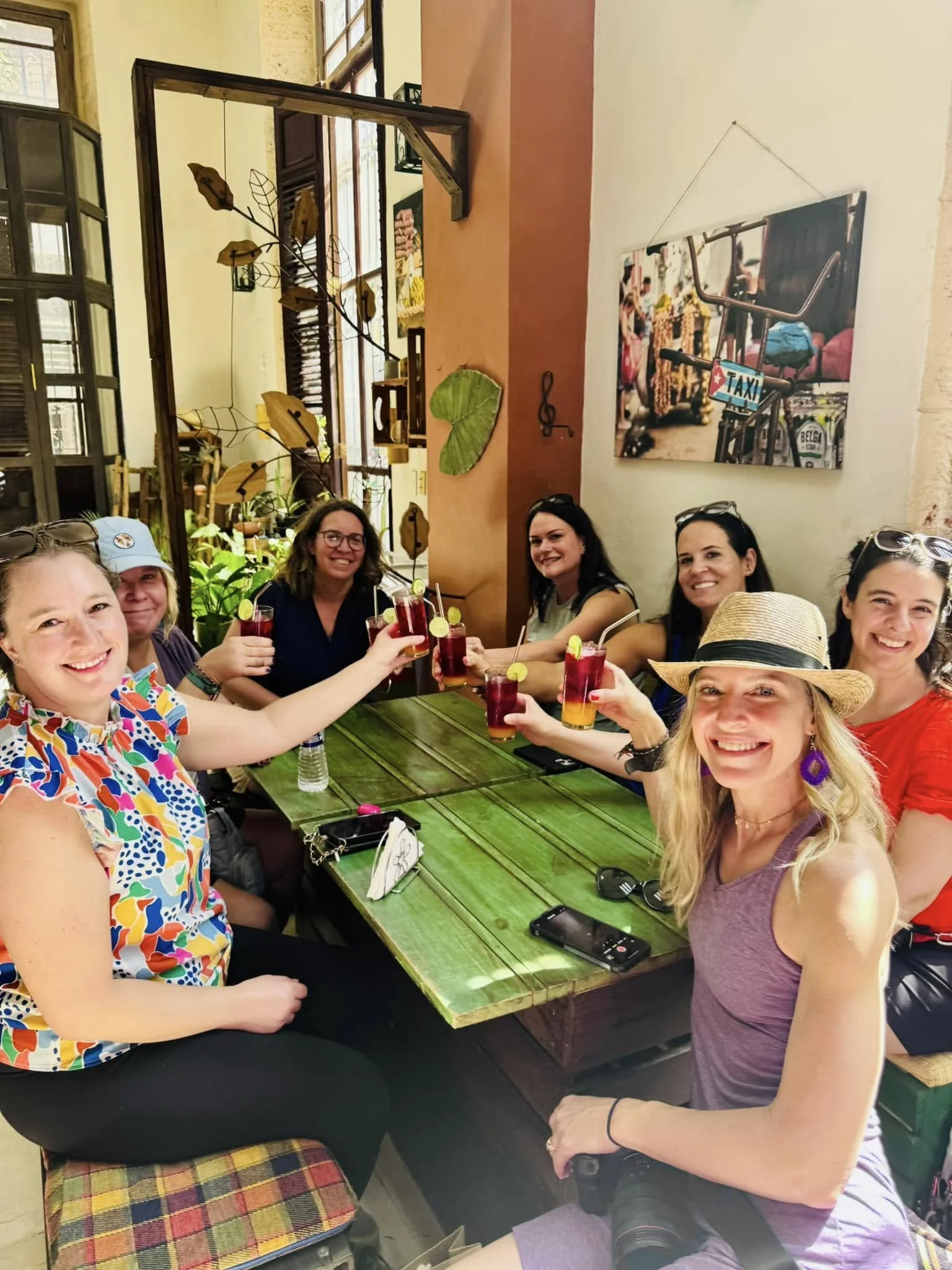 Group of seven women sitting around a green wooden table in a restaurant or cafe, smiling and toasting with colorful drinks garnished with lemon slices, with lively decor and artwork on the walls.