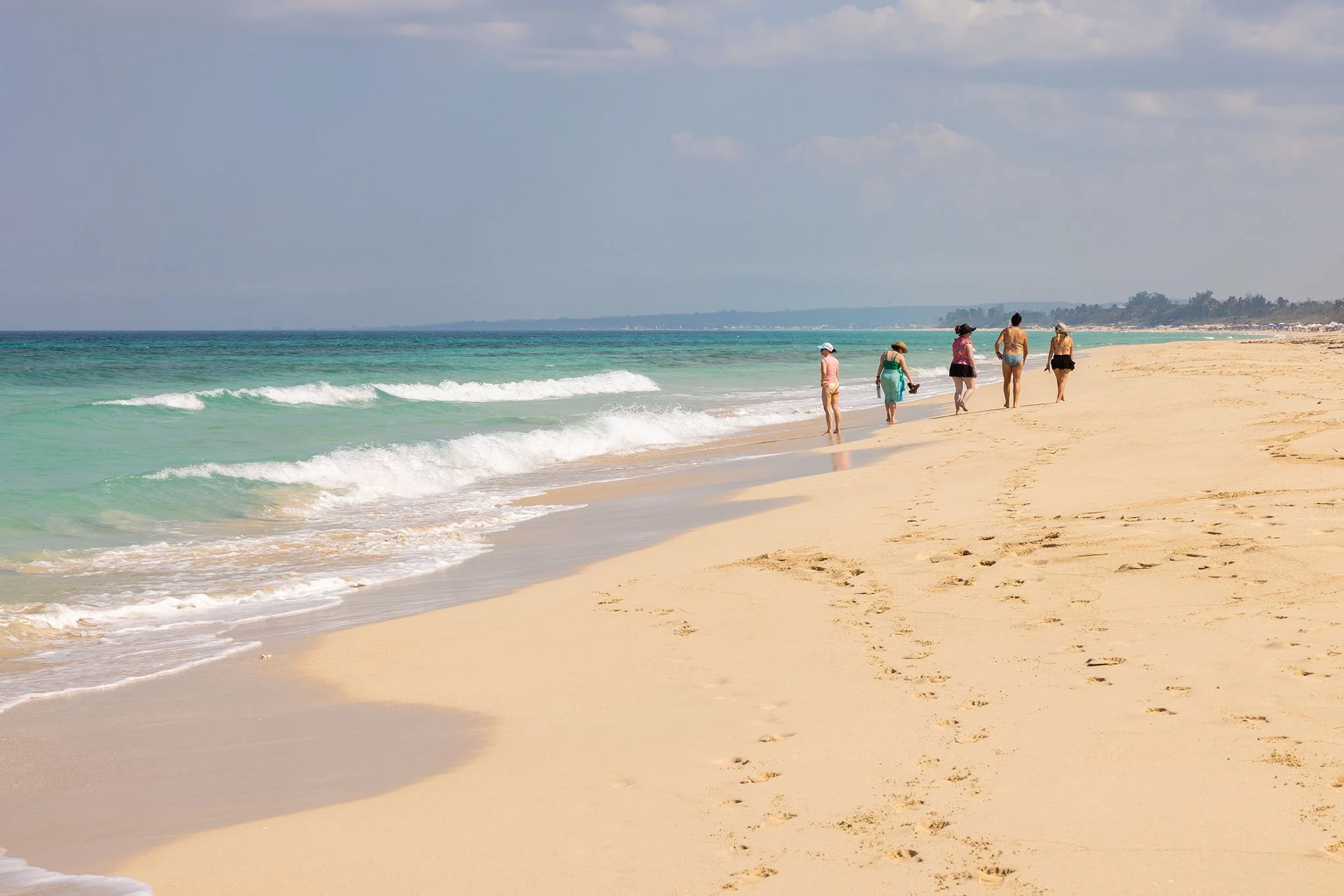 Six women walking along a sandy beach near the water on a sunny day, with gentle waves and distant land visible.