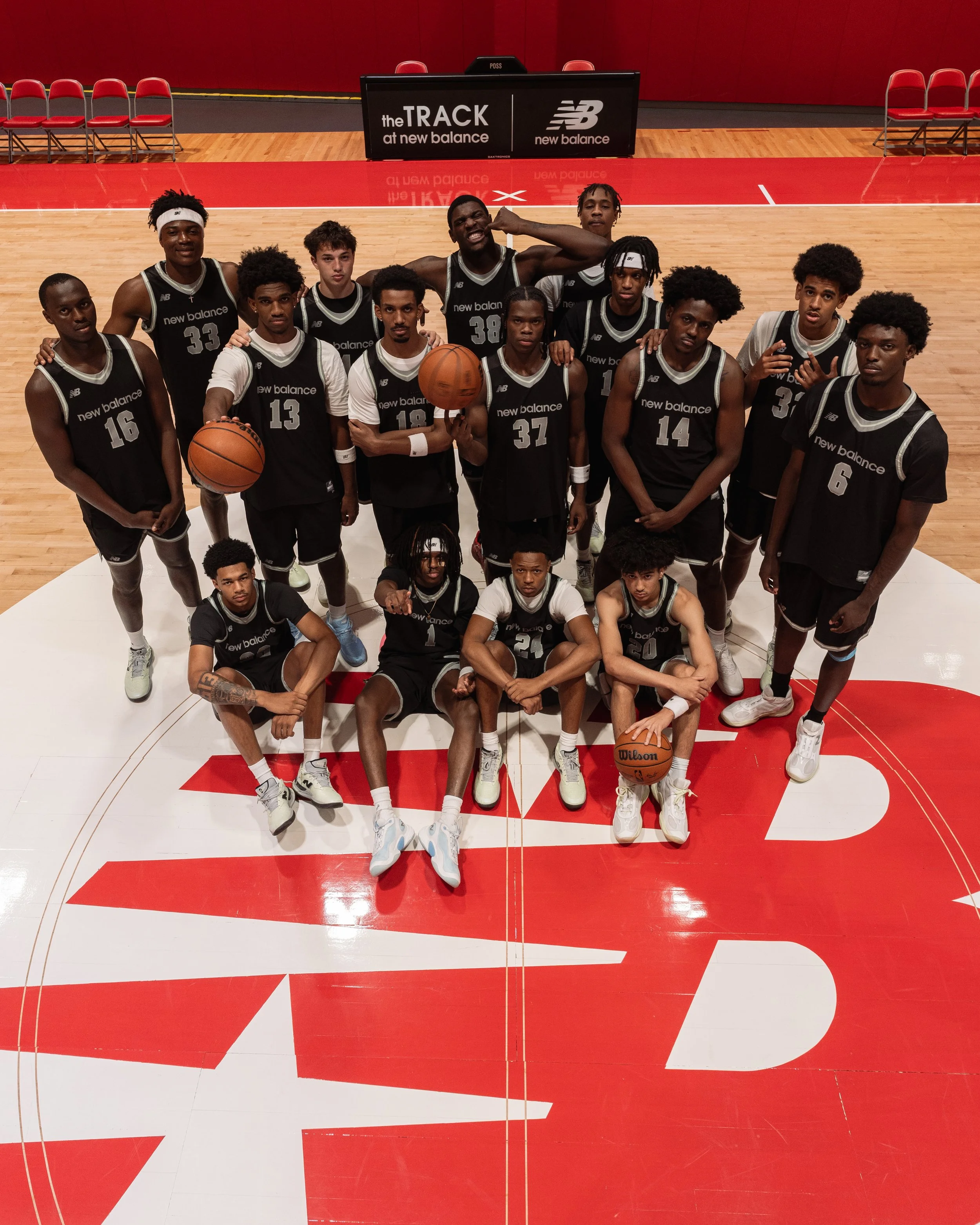 A basketball team posing on the court at New Balance sponsored event. The team consists of 15 young men wearing black and white uniforms, with two players holding basketballs. They are standing and sitting on a red and white basketball court, with a red background and a New Balance promotional banner visible behind them.