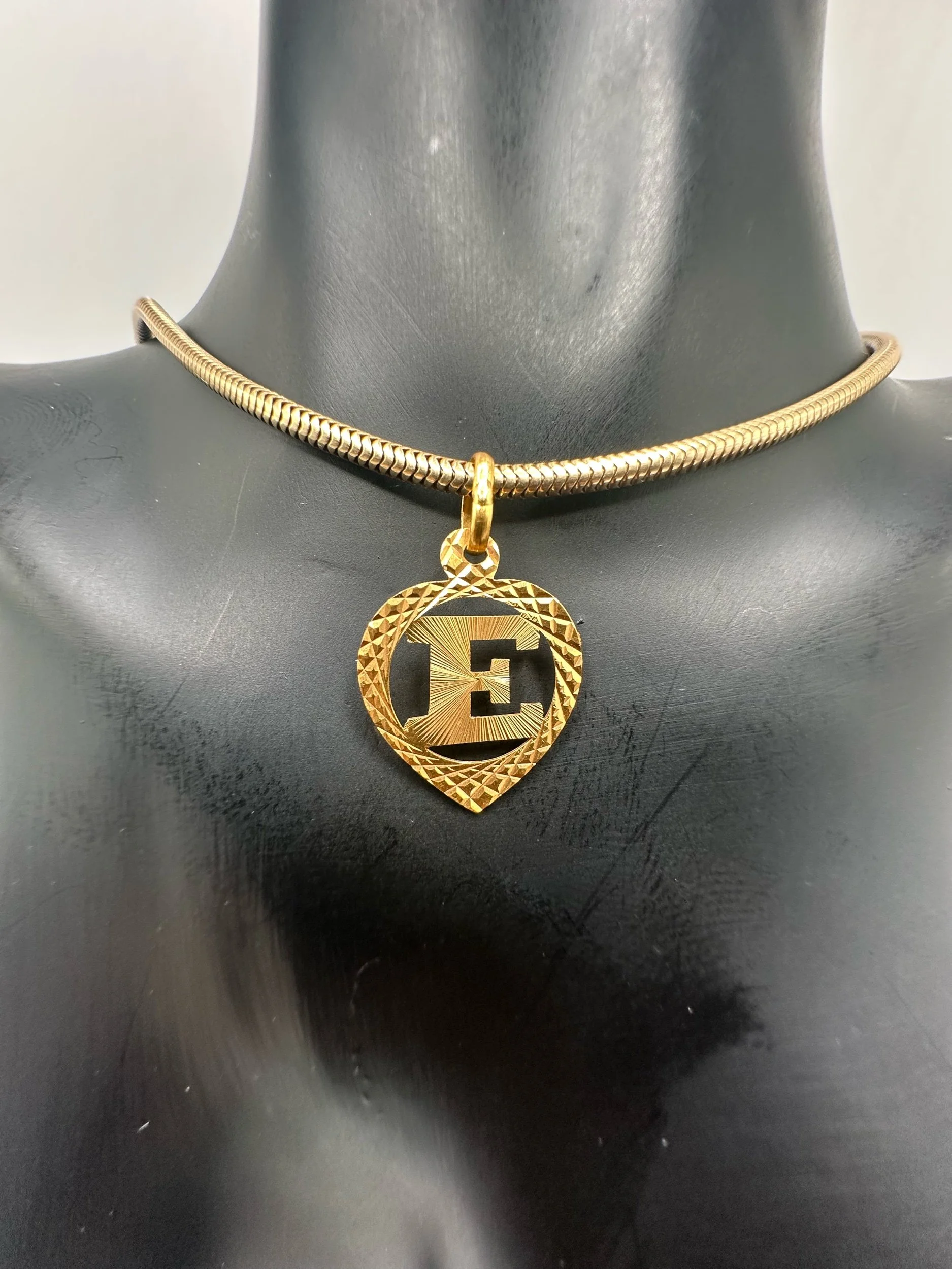 Close-up of a 22k gold letter E charm on 14k gold snake chain on a black mannequin