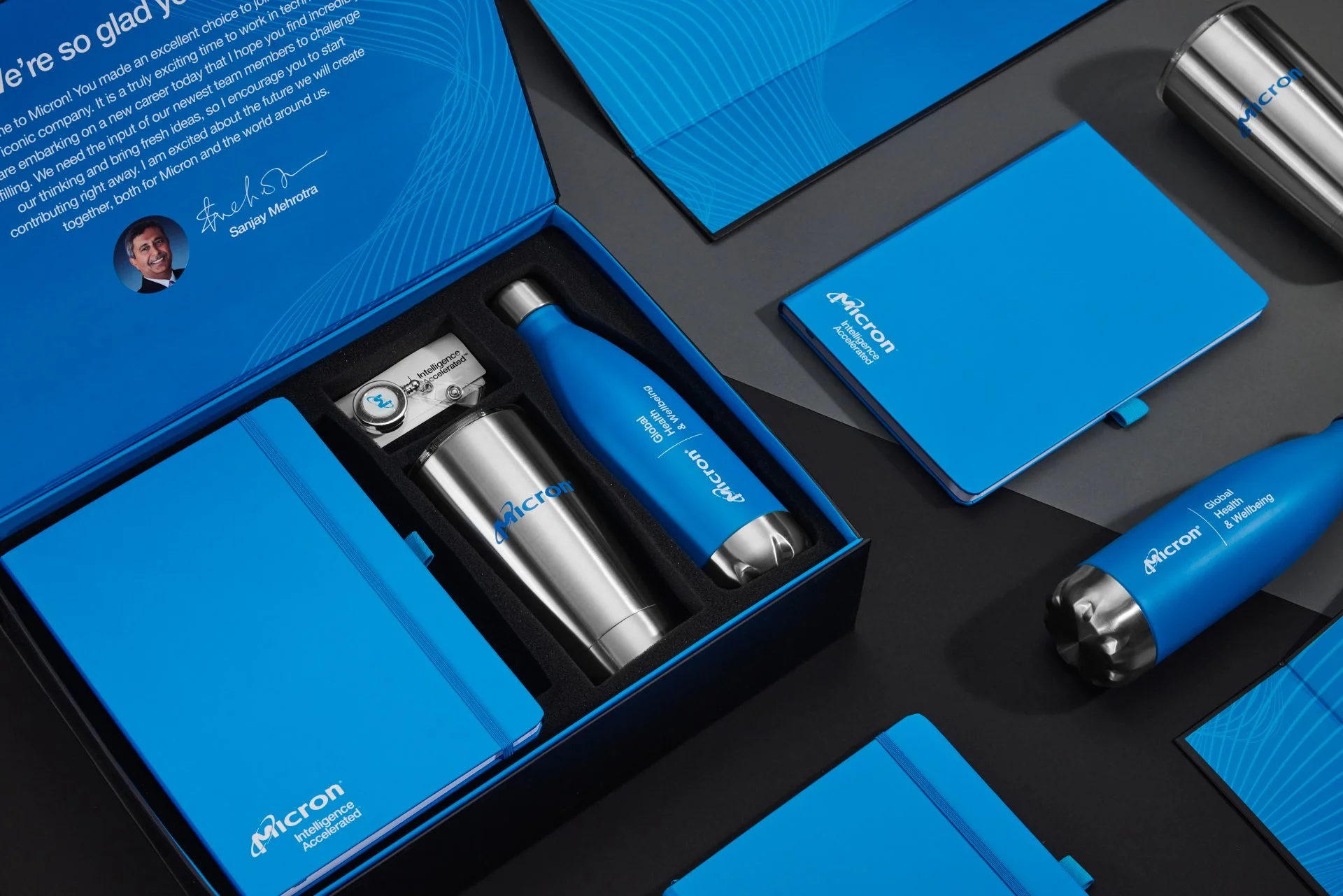 Corporate branded welcome kit with blue packaging, featuring items like a notebook, stainless steel tumbler, water bottle, USB drive, and informational materials with the "Micron" logo.