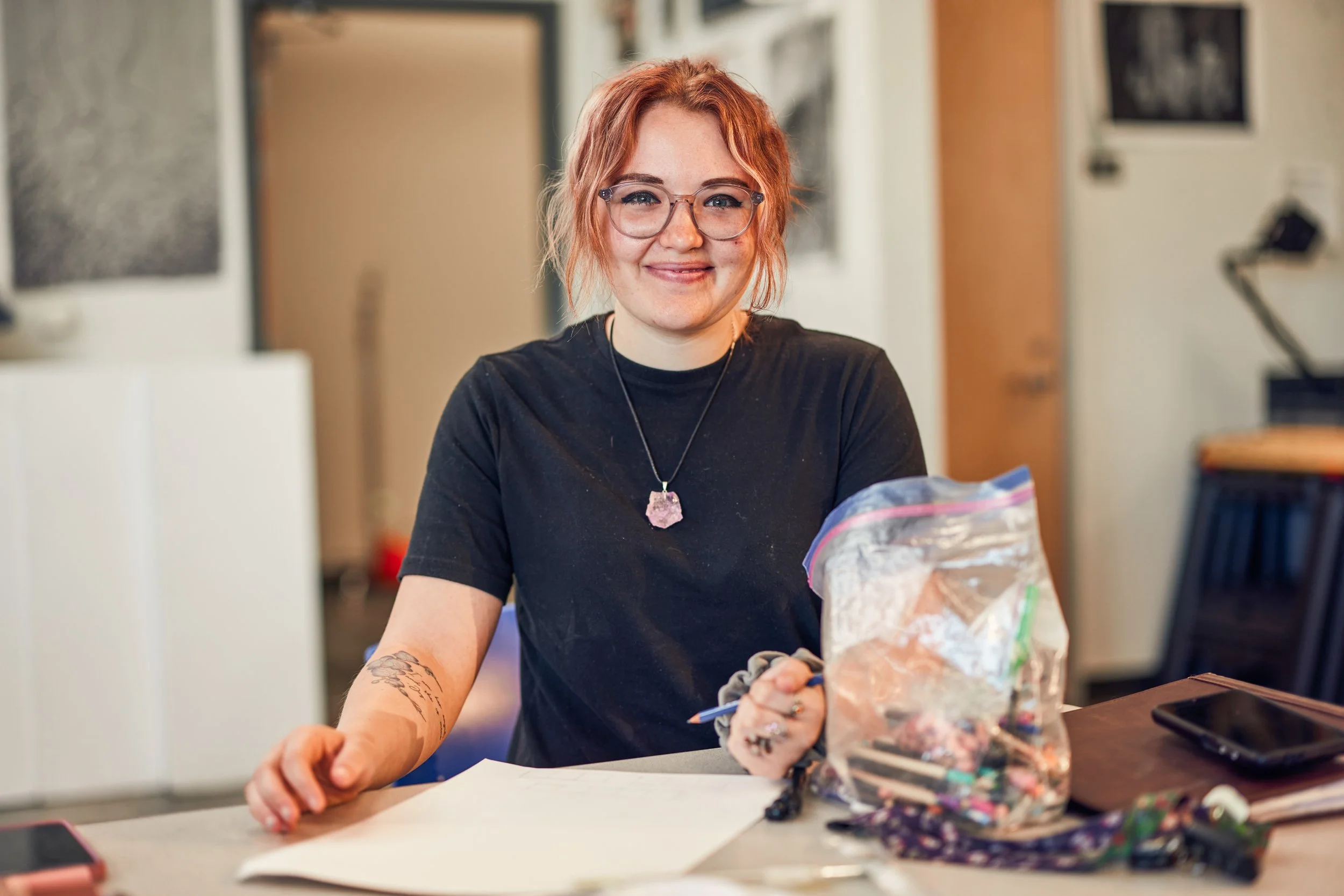 Person with glasses and red hair smiling at a table with art supplies in a studio.