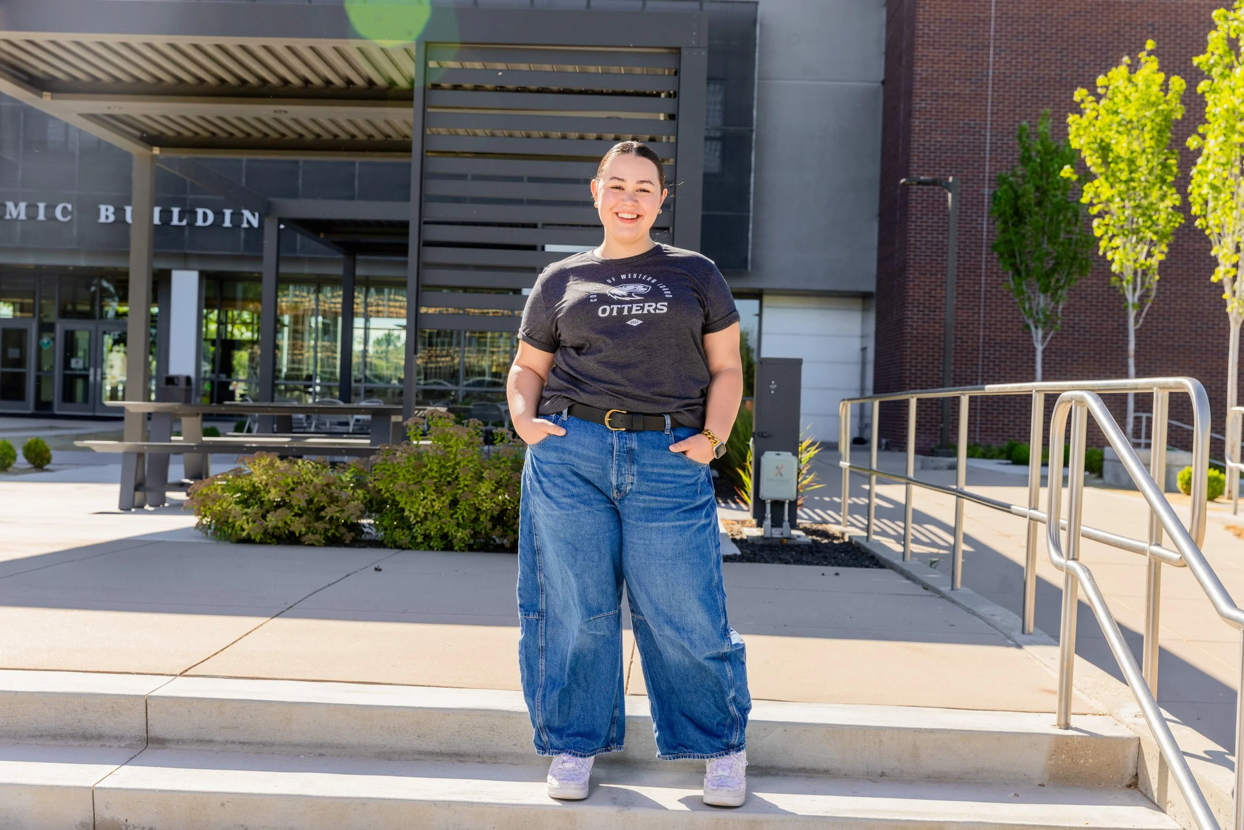 Person wearing a black shirt and blue jeans, standing outside a building with greenery and a metal railing.