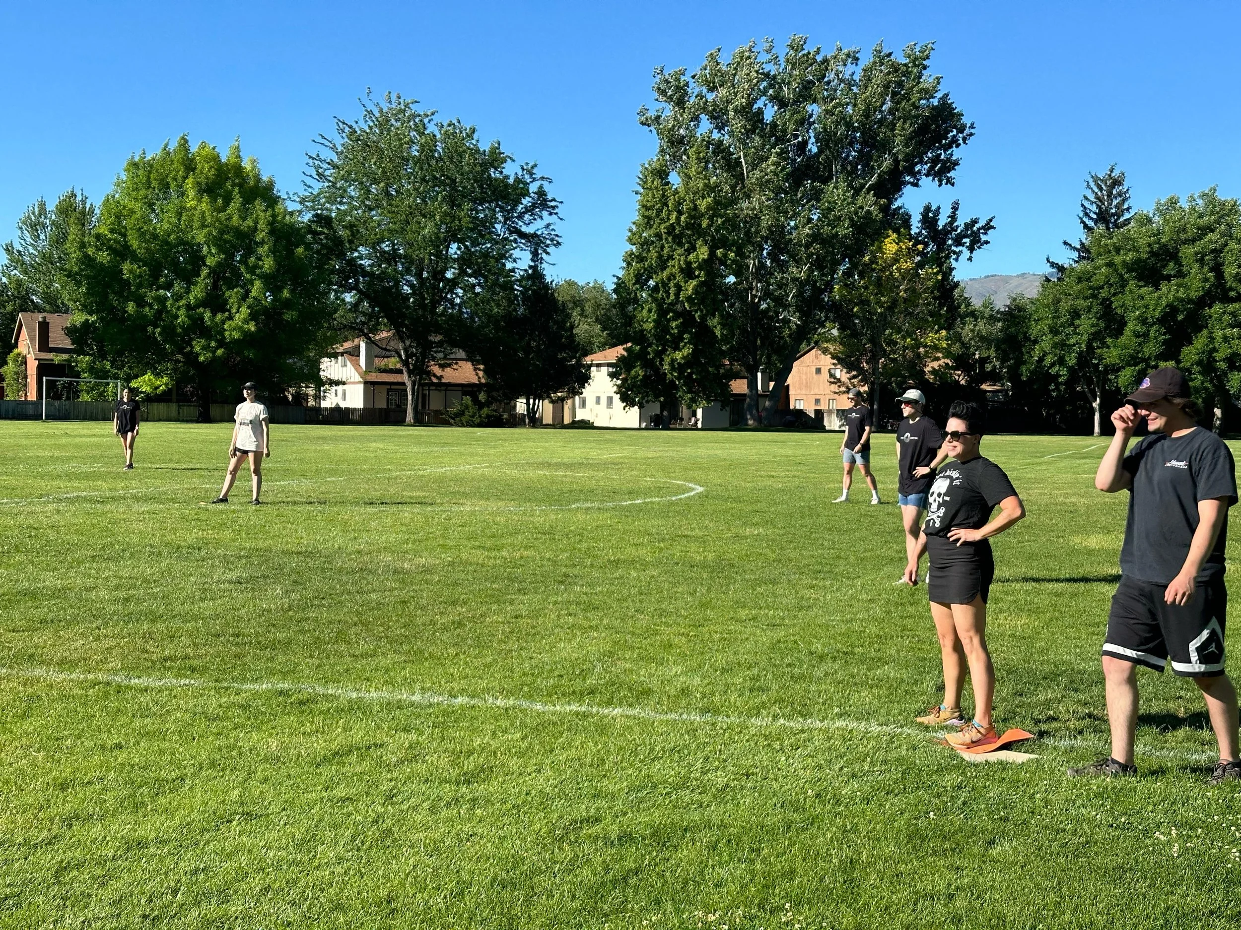 Against team members spread out on a grassy field during a kickball game, with a few players standing near a base and others in the outfield under a clear blue sky.