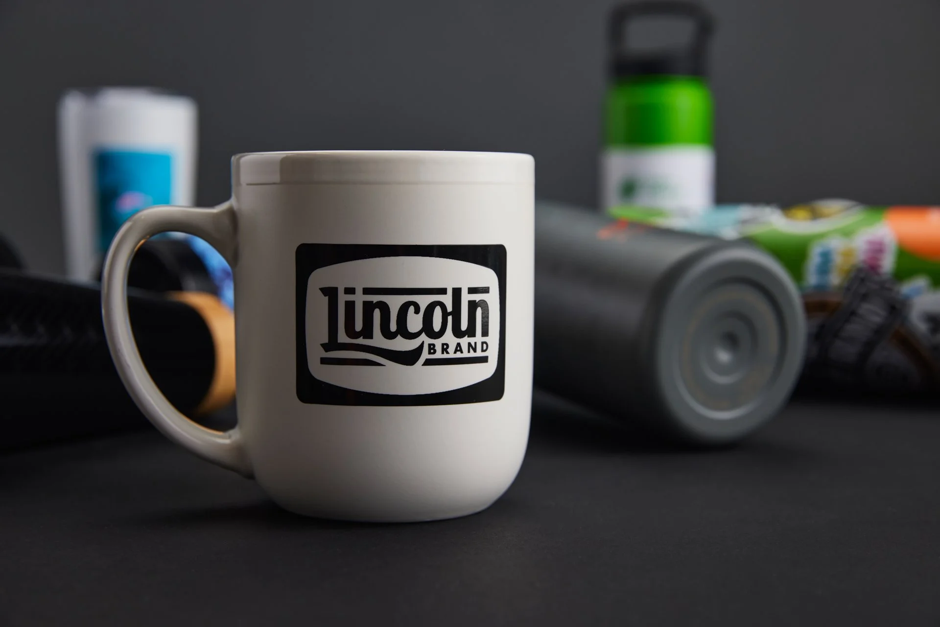 White coffee mug with "Lincoln Brand" logo, surrounded by blurred objects including a green bottle and colorful packaging, on a dark surface.
