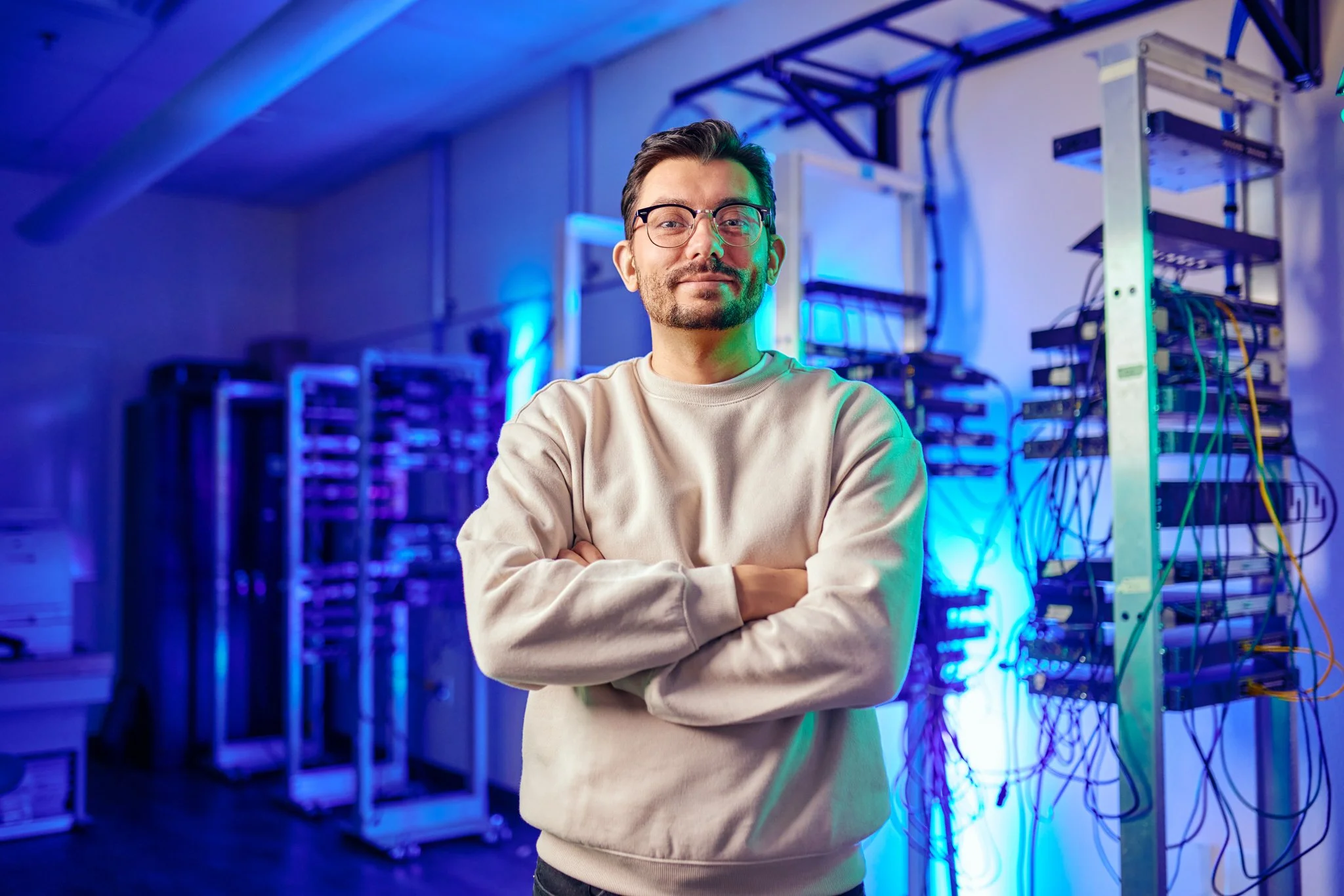 Person in glasses and a sweater standing confidently in a data center with server racks.